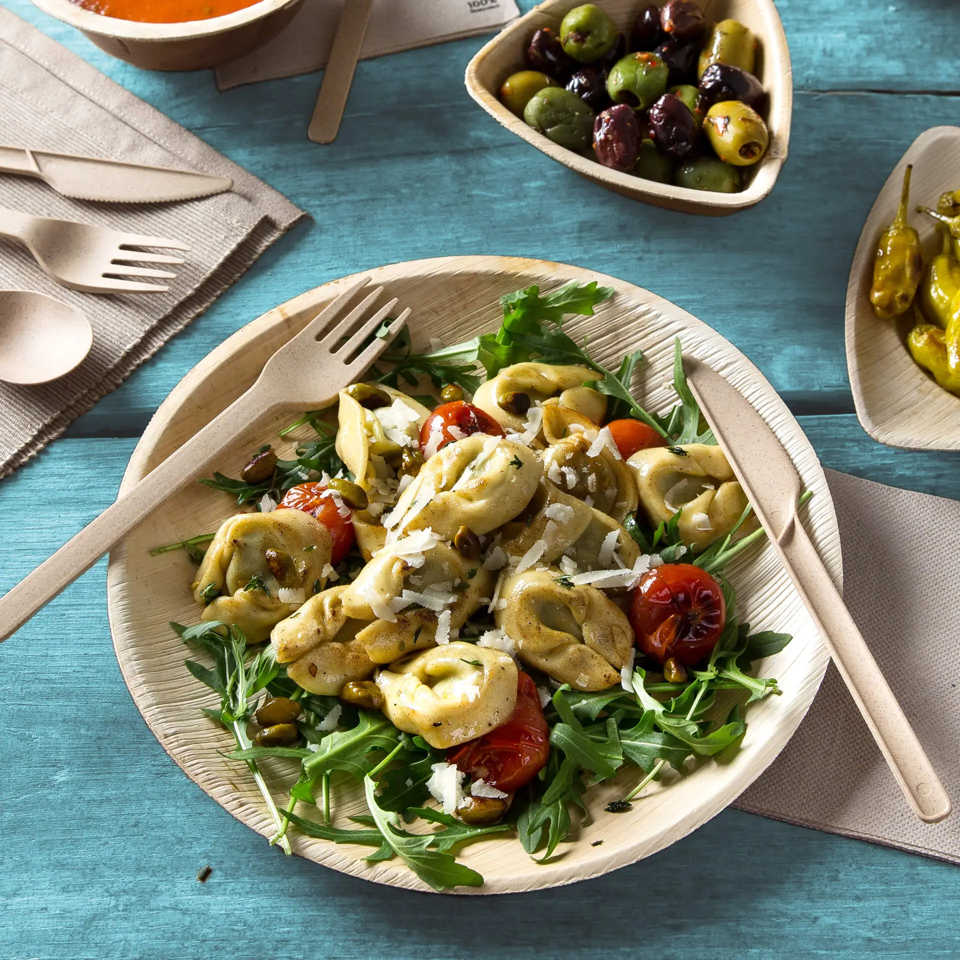 Un piatto rotondo in foglia di palma con insalata di pasta, pomodori e rucola; accanto utensili in legno e ciotole di olive e peperoncini.