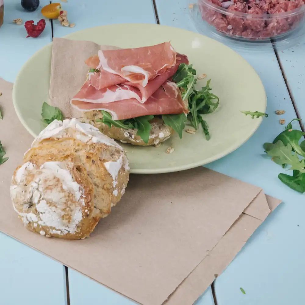 Fette di prosciutto su un piatto verde con rucola su un panino marrone; un panino rotondo e sfogliato è vicino.