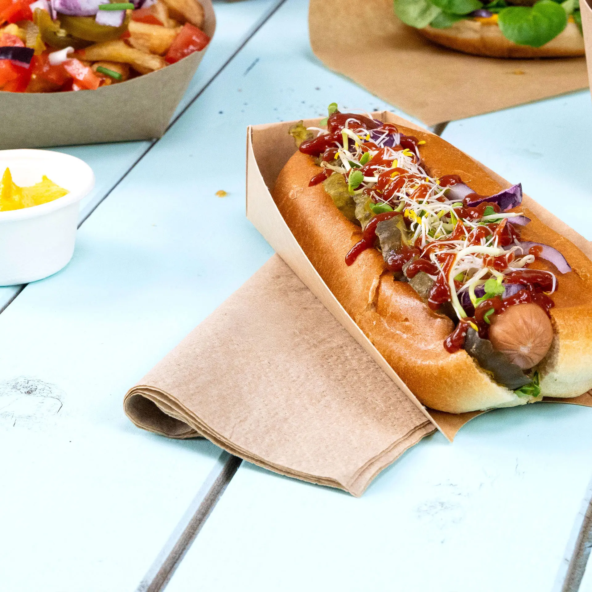Hot dog in un support di carta, guarnito con ketchup, senape, cetriolini e germogli, con un tovagliolo accanto su un tavolo blu.