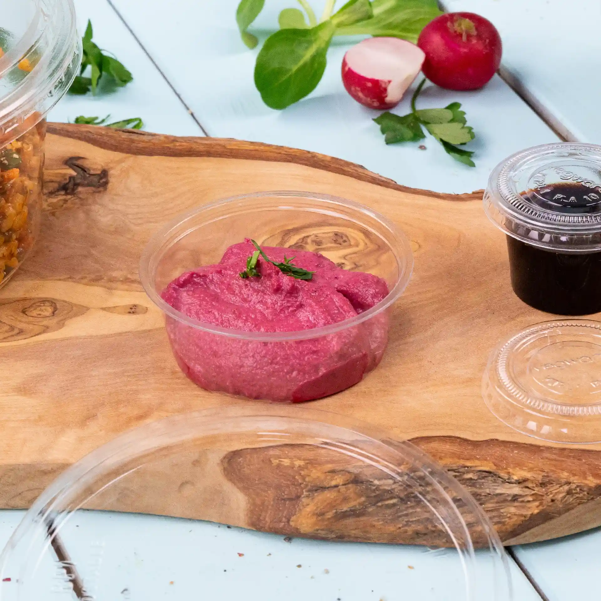 Vasetto per dip trasparente rotondo con dip rosa, guarnito con una piccola erba verde, su una superficie di legno con verdure.