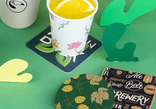 Pile di sottobicchieri su una superficie in legno; alcuni presentano un logo con un uccello, altri un design con birra e testo.