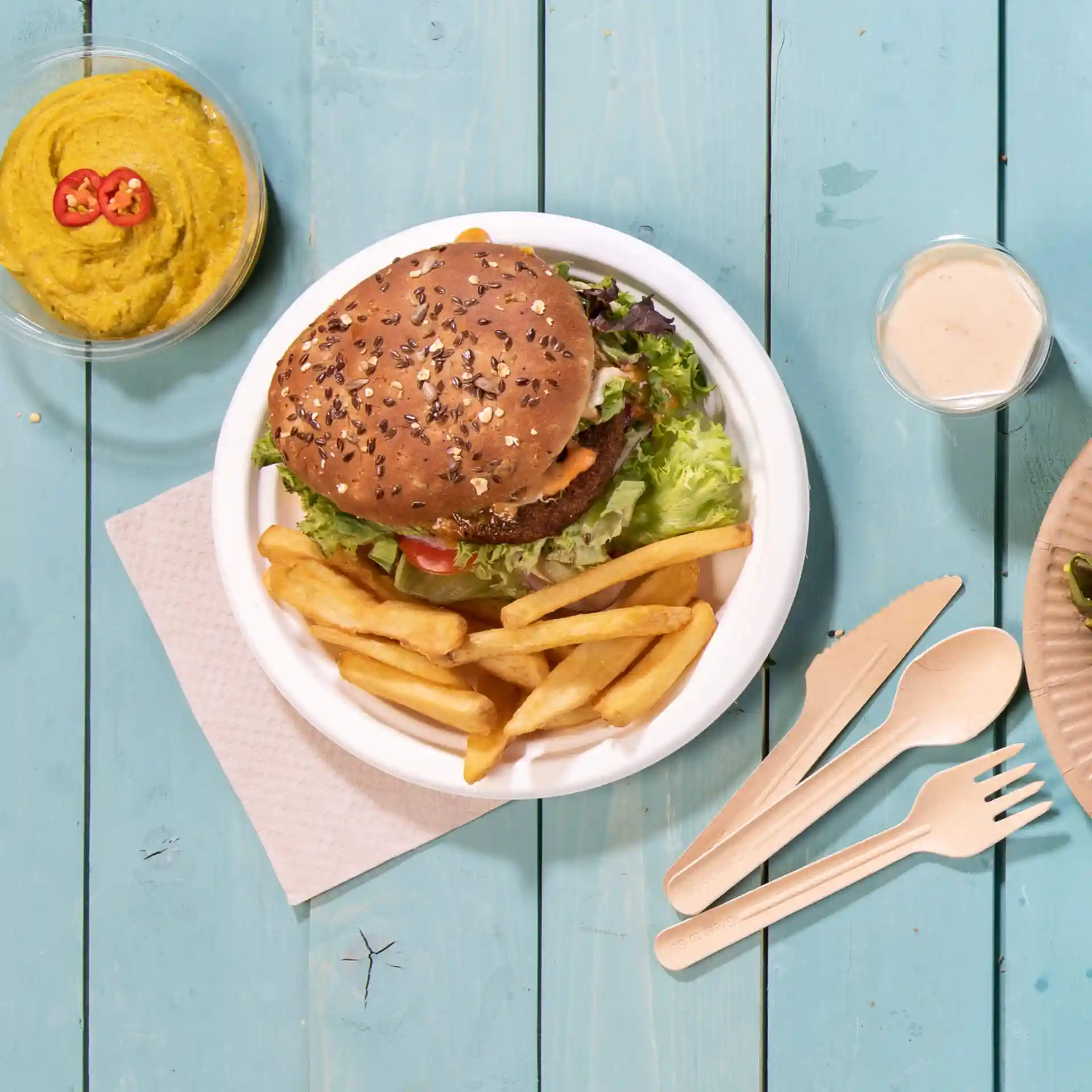 Immagine di un piatto rotondo bianco in canna da zucchero con un hamburger, patatine e una ciotola di hummus su una superficie in legno blu.