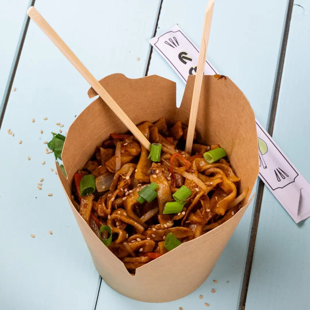 Contenitore di carta marrone riempito di noodles saltati, guarnito con cipollotti e semi di sesamo, con bacchette inserite.