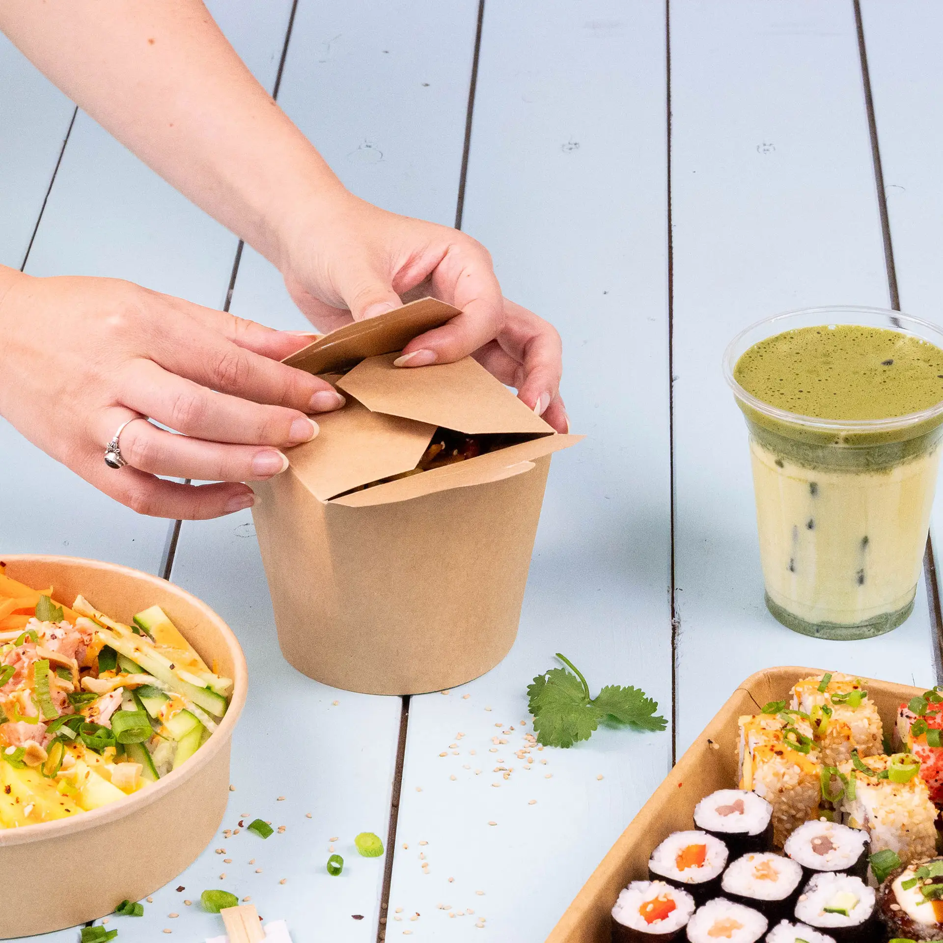 Una scatola di noodle marrone riempita di noodle fritti, guarnita con peperoncini rossi affettati e coriandolo, su un tappetino di bambù con altri contenitori alimentari.