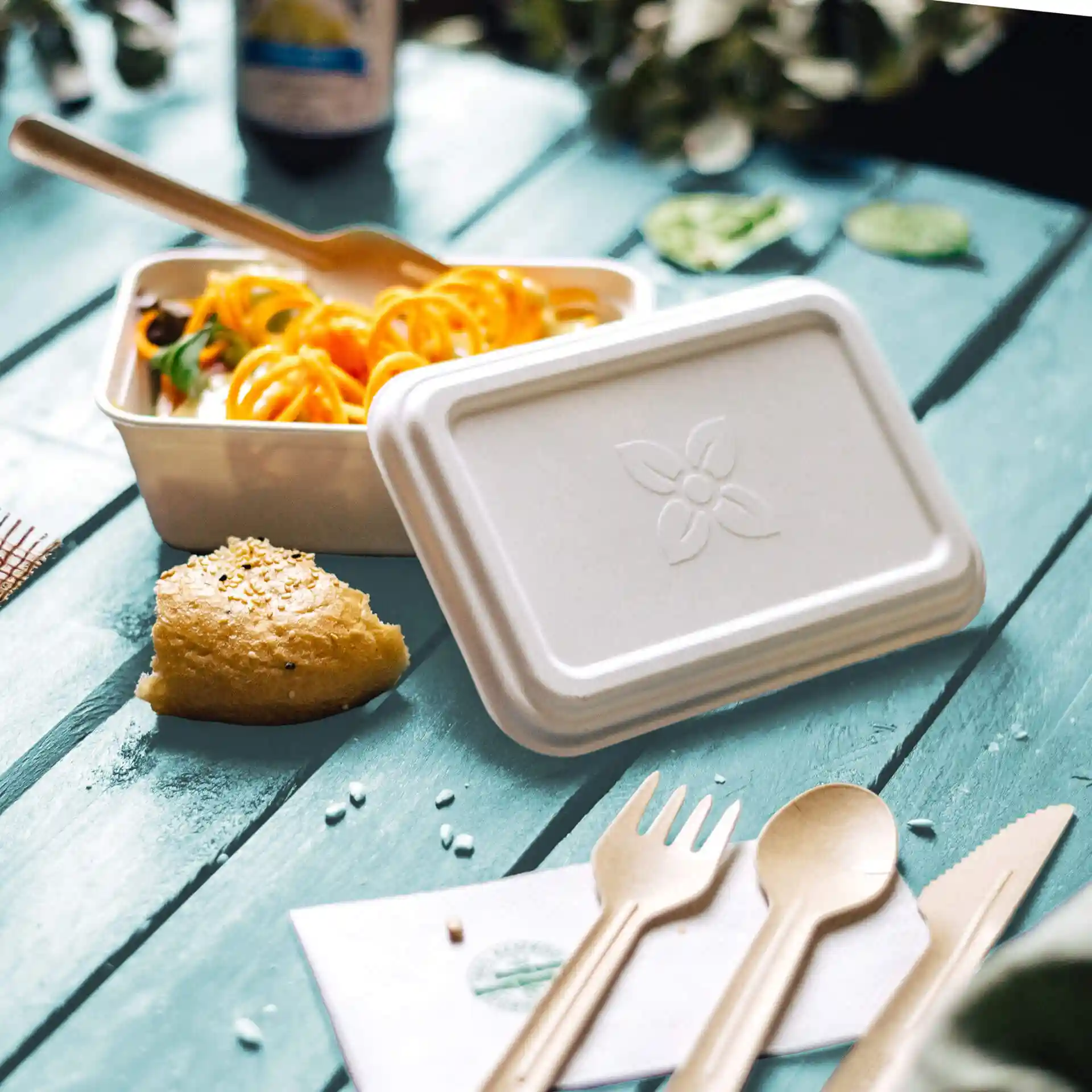 Contenitore rettangolare bianco in canna da zucchero con coperchio, forchetta, cucchiaio e un pezzo di pane su una superficie in legno blu.