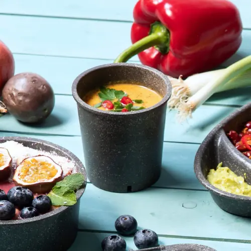 Tazza di zuppa riutilizzabile riempita di zuppa e guarnita con erbe, circondata da verdure e frutta fresca su una superficie blu.