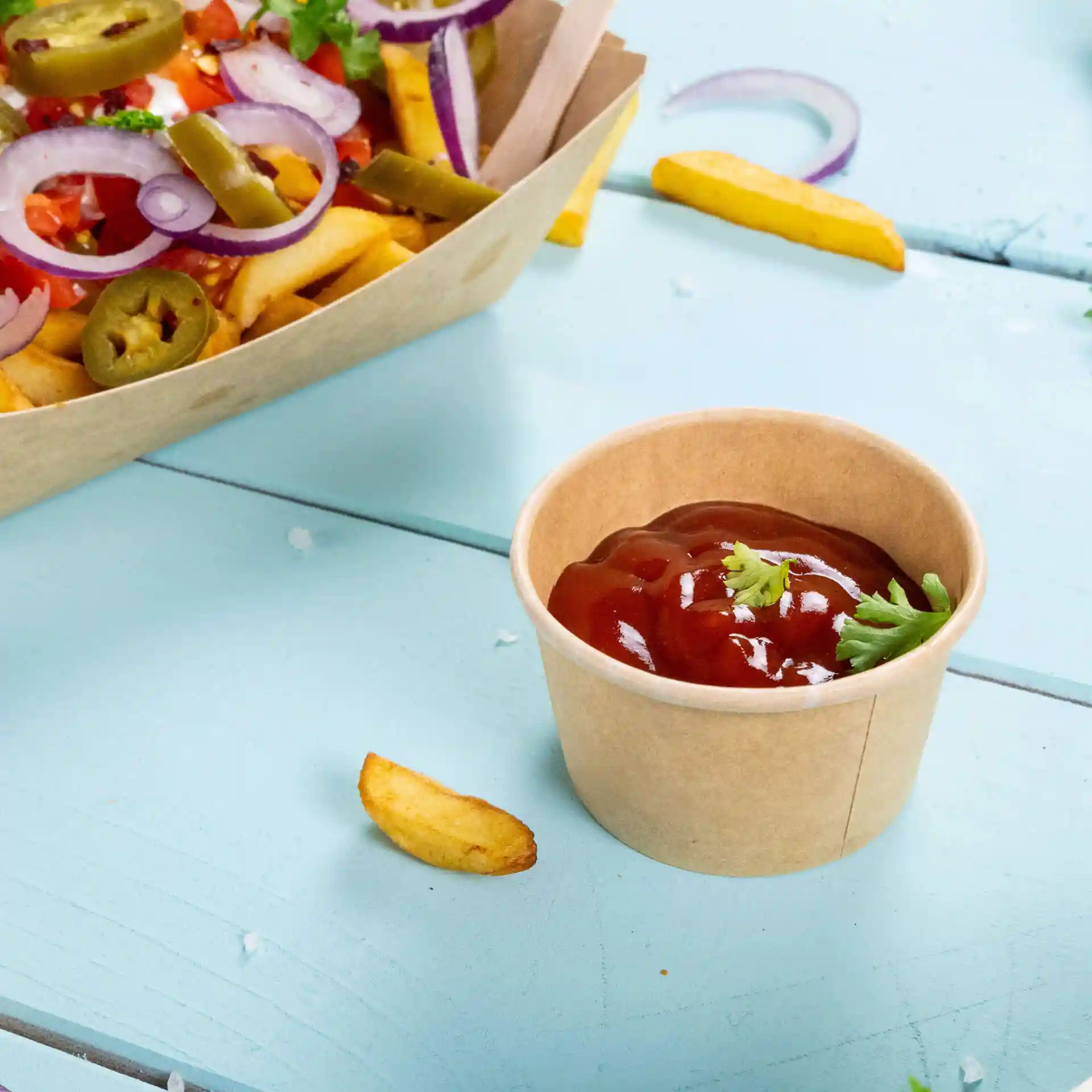 Contenitore rotondo in cartone marrone con ketchup e una guarnizione di coriandolo, posizionato su una superficie in legno blu.