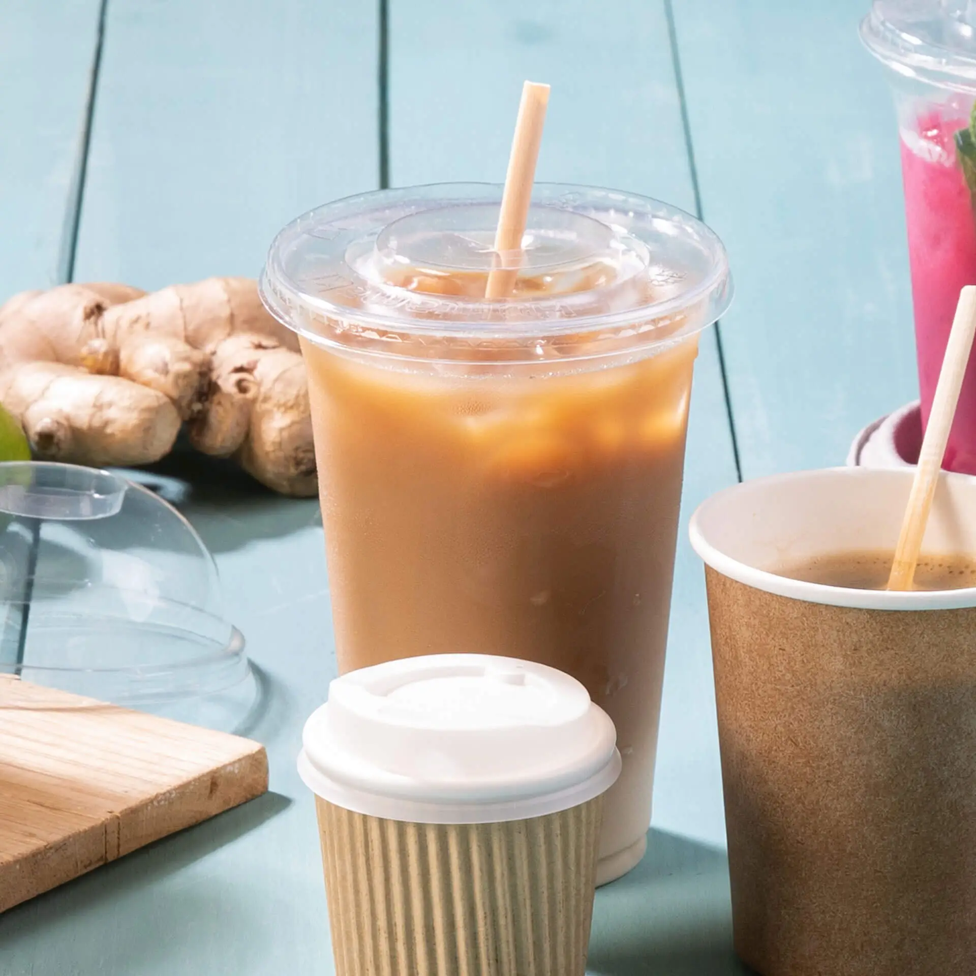 Un bicchiere di plastica trasparente con bevanda ghiacciata e cannuccia; un bicchiere di carta più piccolo con coperchio davanti; radice di zenzero sullo sfondo.