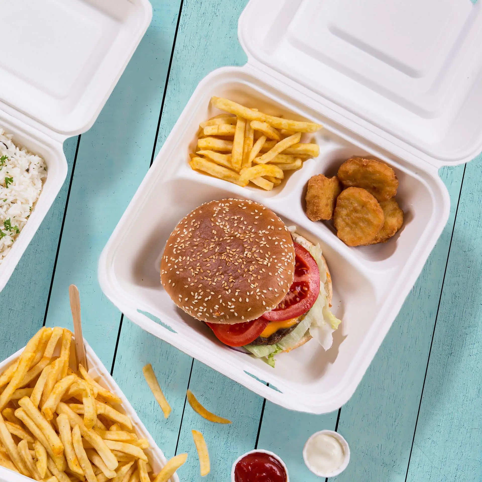 Una scatola per menu biodegradabile a tre scomparti contenente un hamburger ai semi di sesamo, patatine e nuggets di pollo su un tavolo di legno blu.