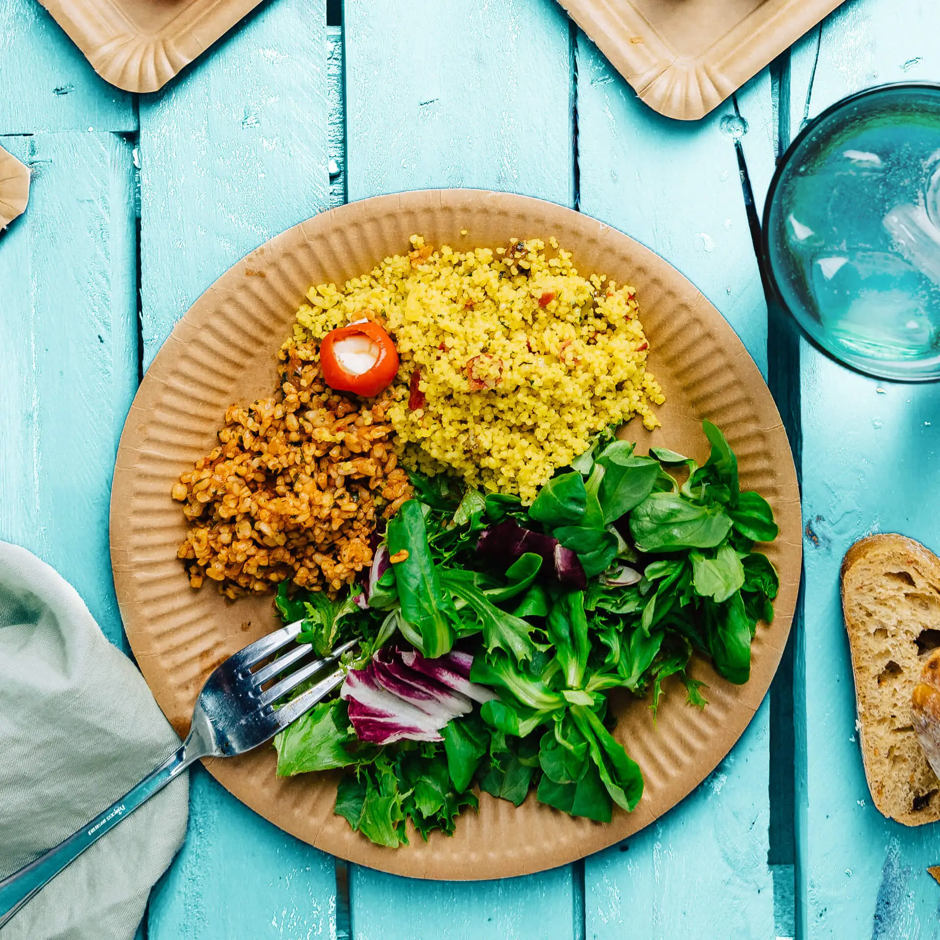 Piatto rotondo in cartone marrone con couscous giallo, cereali marroni e verdure miste; forchetta appoggiata sull'insalata.