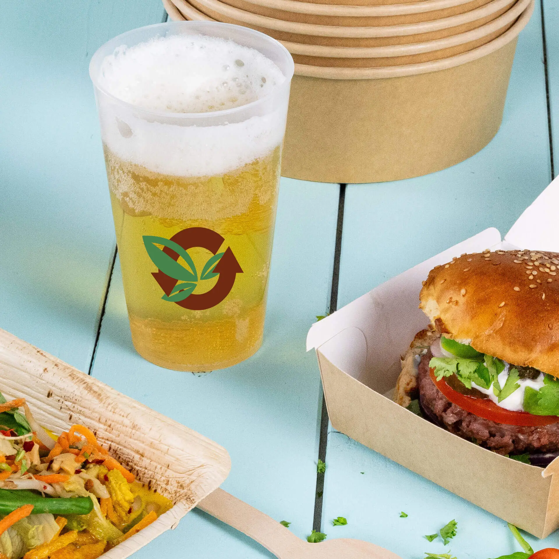 Bicchiere riutilizzabile trasparente con logo, pieno di birra, accanto a un hamburger e un'insalata su una superficie blu.