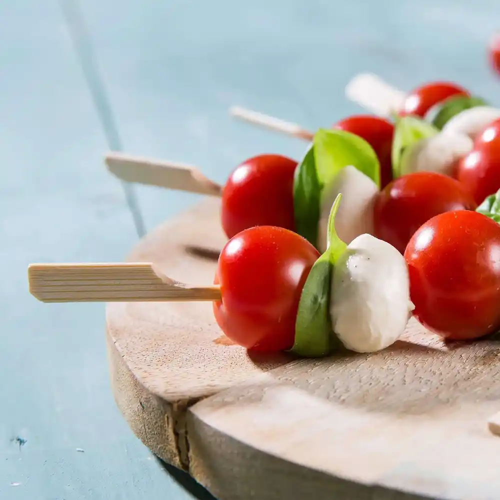 Spiedini con pomodorini, foglie di basilico e palline di mozzarella disposti su un tagliere di legno.