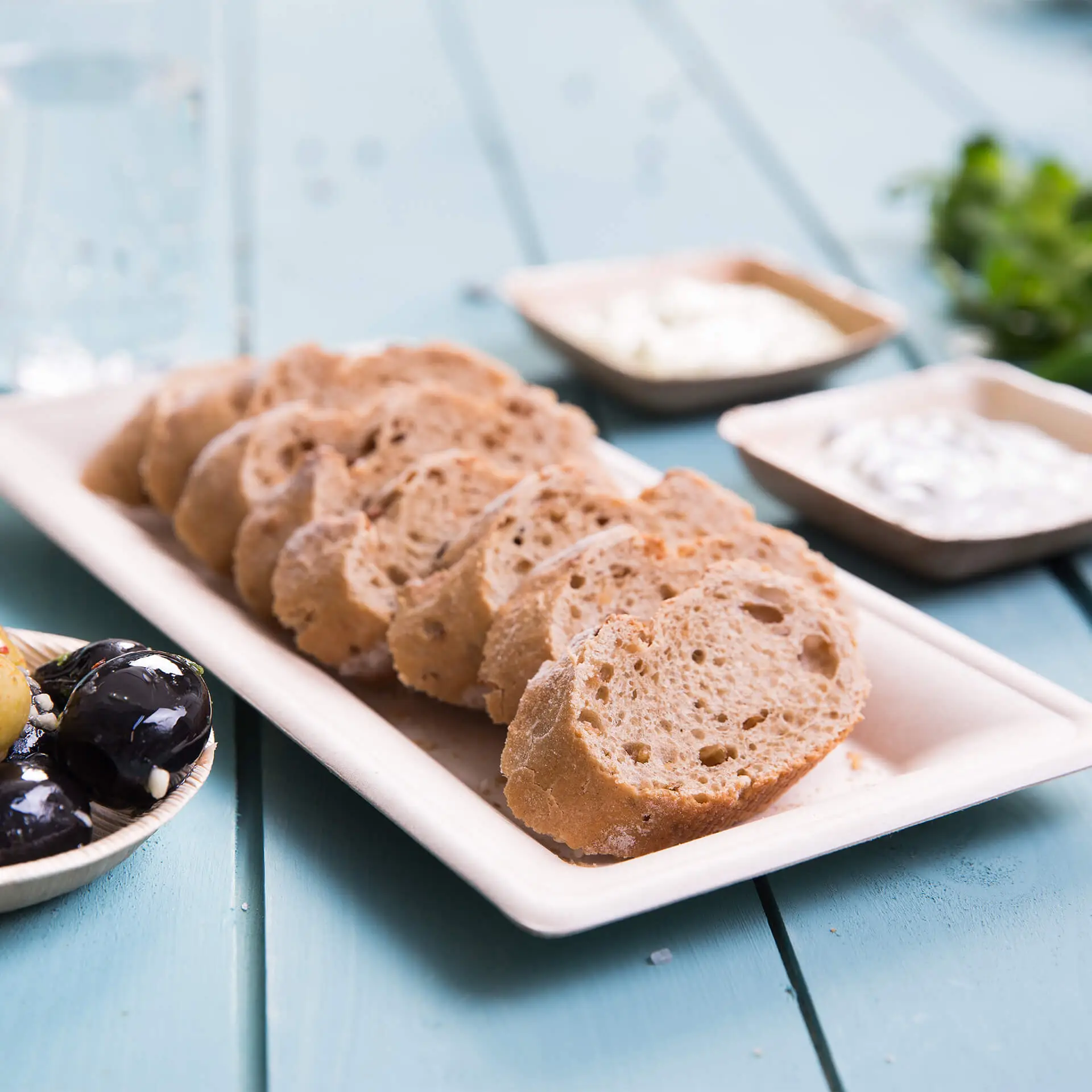 Pane affettato su un piatto rettangolare biodegradabile in canna da zucchero con condimenti e olive sullo sfondo.