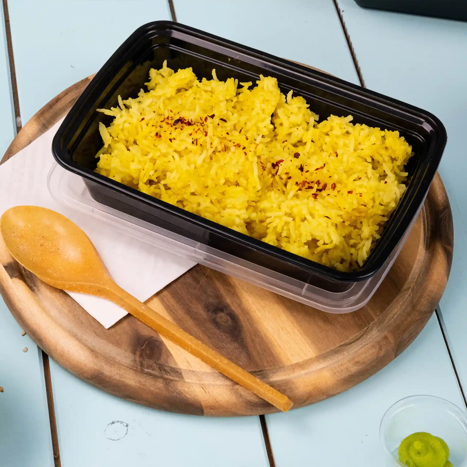 Contenitore rettangolare riutilizzabile nero con riso giallo all'interno, cucchiaio di legno accanto, su un tagliere rotondo.