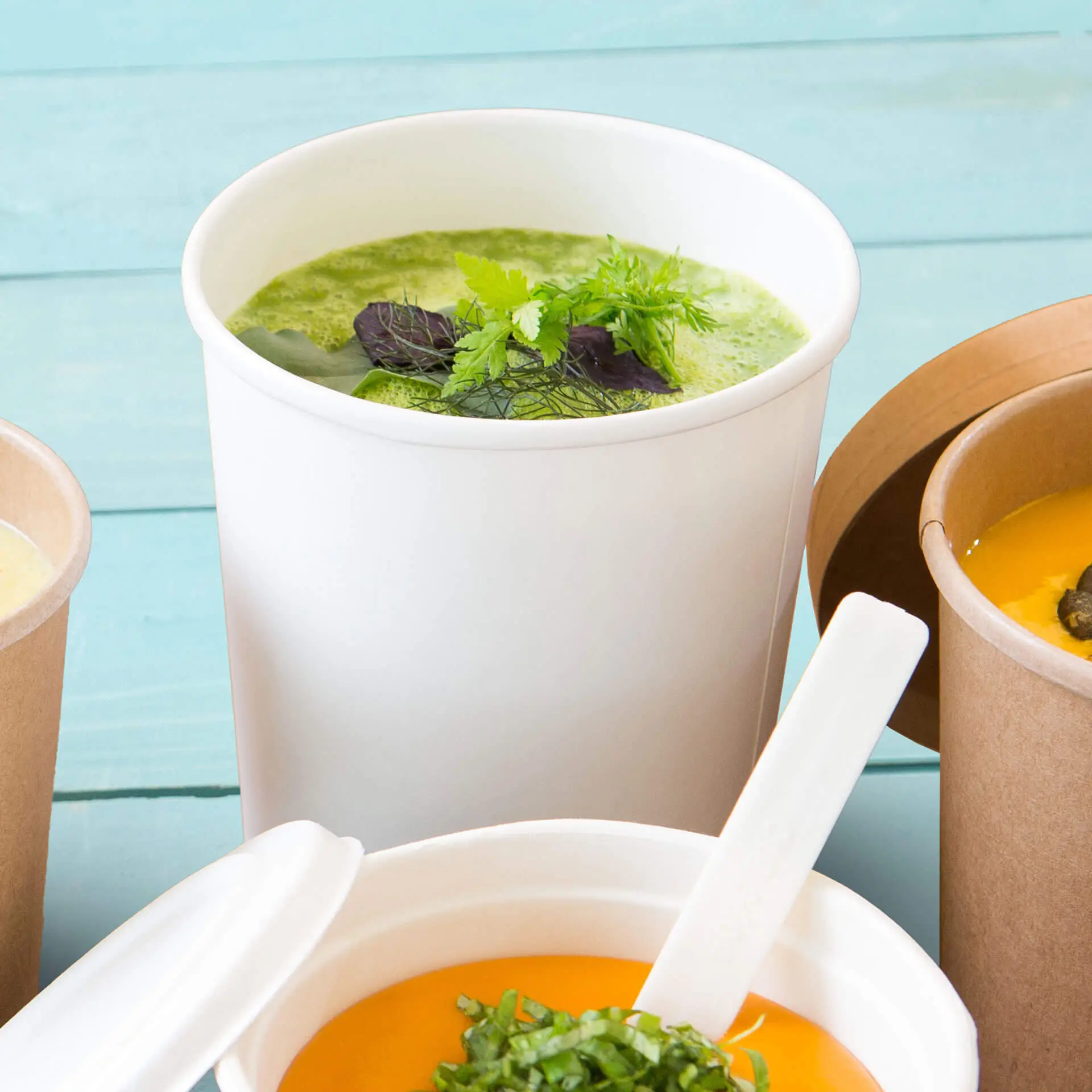 Tazza per zuppa biodegradabile bianca con zuppa verde e erbe, posizionata con altri contenitori per zuppa su uno sfondo turchese.