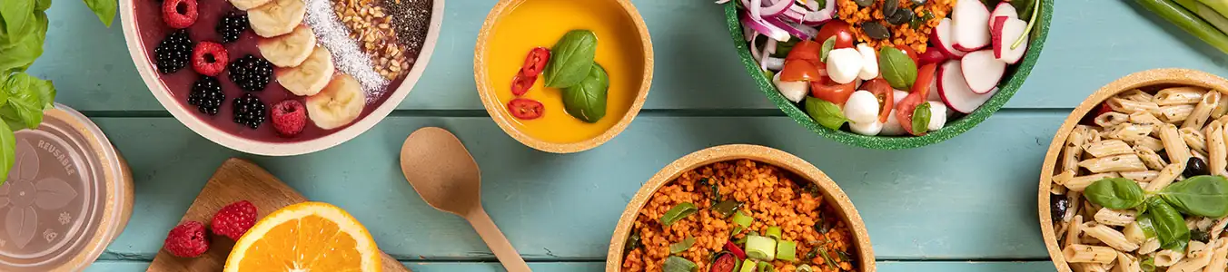 Ciotole colorate piene di vari alimenti, tra cui frutta, pasta, insalata e cereali, disposte su un tavolo di legno.