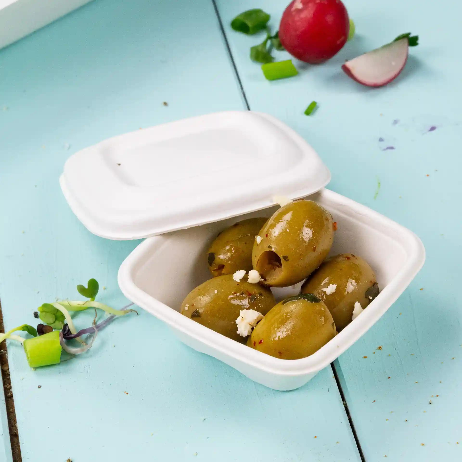 Contenitore con diverse olive verdi all'interno, posato su una superficie azzurro chiaro, con un coperchio bianco parzialmente chiuso.