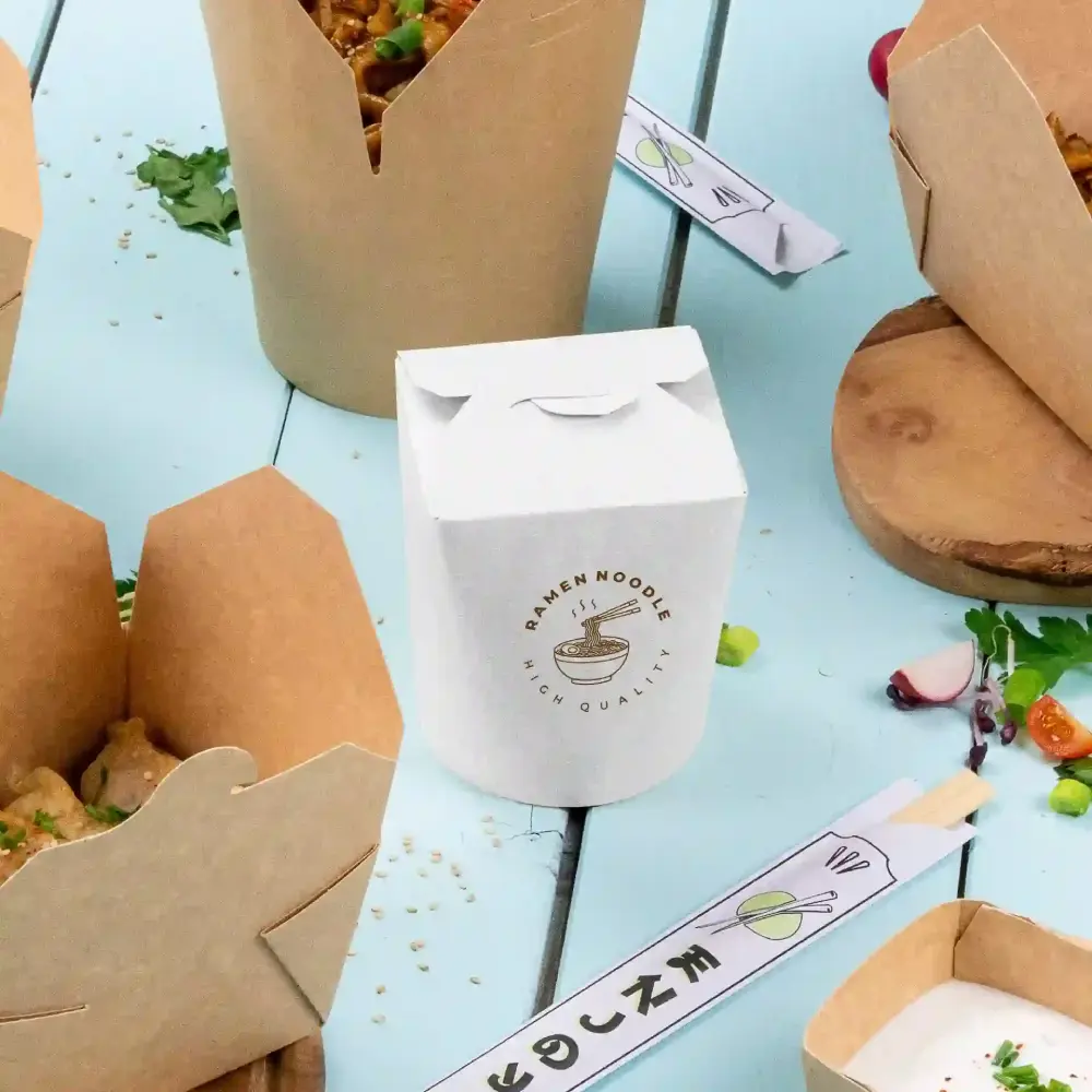 Scatola da asporto bianca etichettata "Ramen Noodle di alta qualità" accanto a scatole da asporto marroni su una superficie blu.