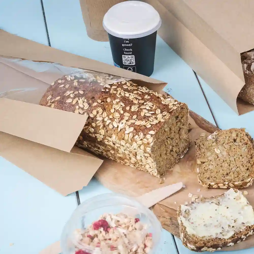 Pane d'avena affettato su un tagliere di legno, con un bicchiere e un contenitore di granola vicino.