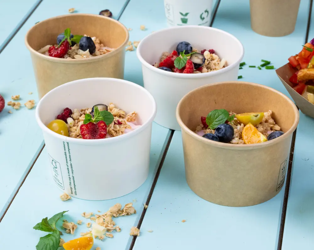 Quattro contenitori per alimenti pieni di granola e frutta fresca, comprese bacche e agrumi, su una superficie in legno blu.