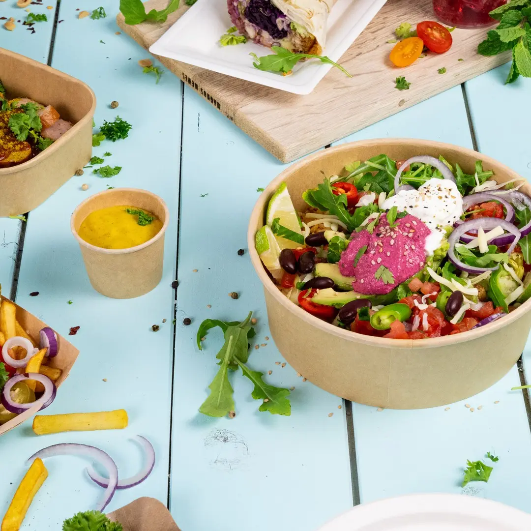 Ciotola di insalata con verdure miste, lime e salse, circondata da contenitori più piccoli di salsa, patatine e erbe su una superficie blu.