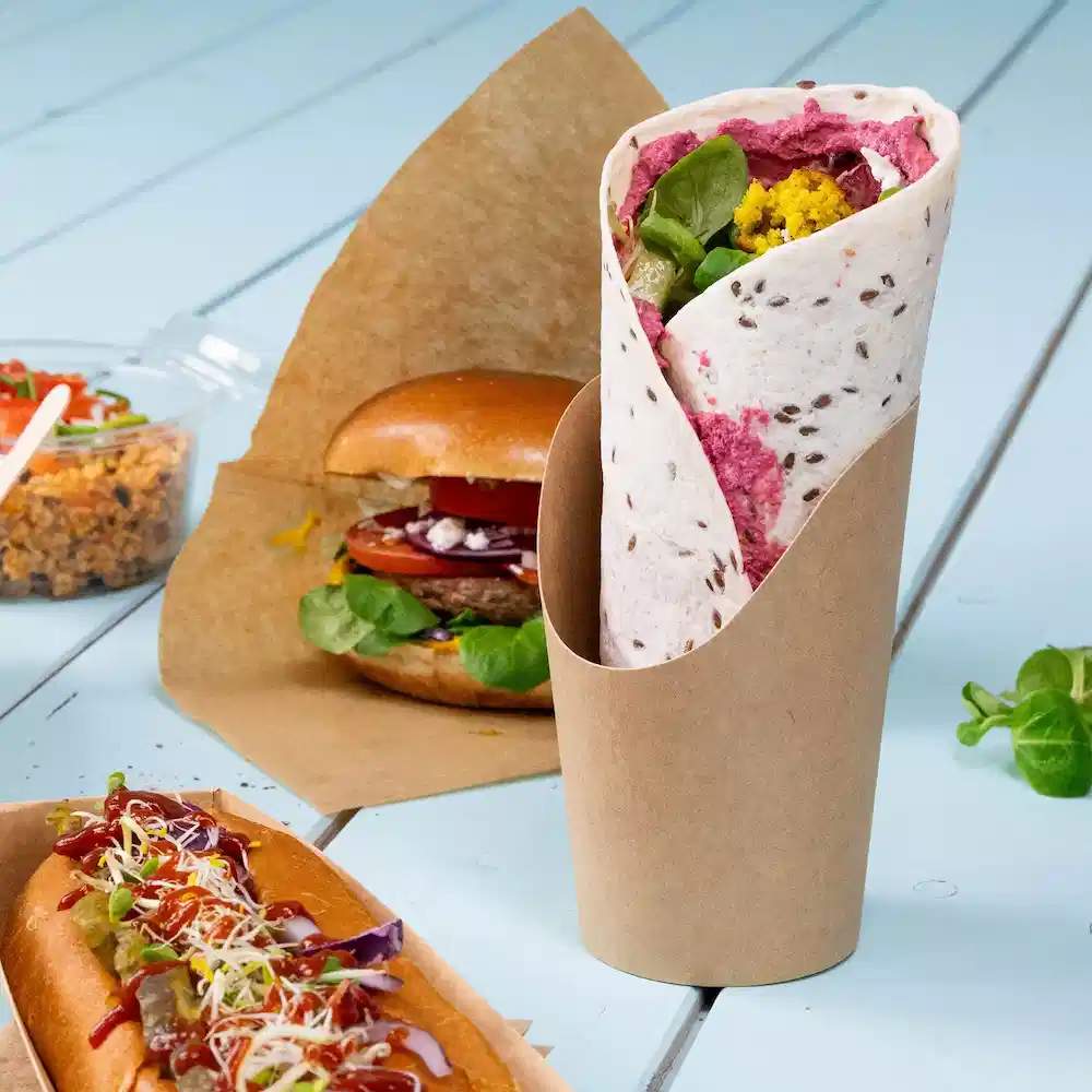 Wrap farcito con verdure e crema, servito in un supporto di carta marrone, con un hamburger e un hot dog sullo sfondo.