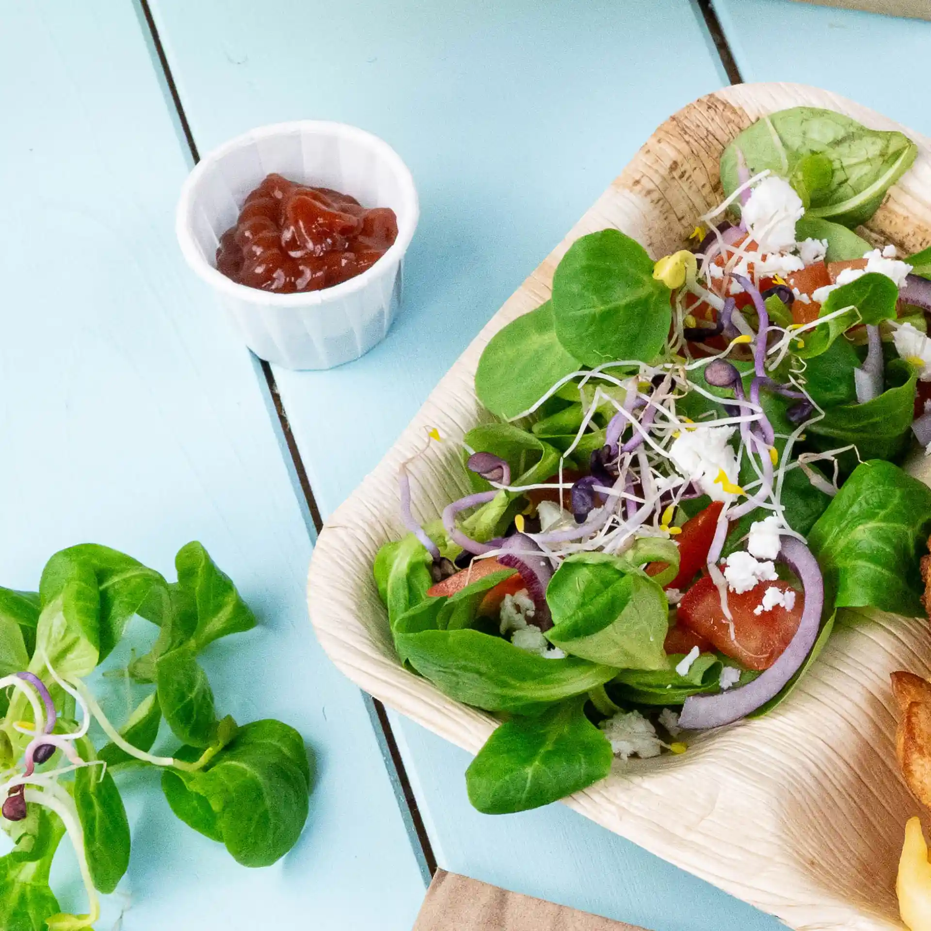 Insalata con misto di verdure, pomodori, cipolle e germogli in una ciotola di foglia di palma, accompagnata da una piccola tazza bianca di ketchup.
