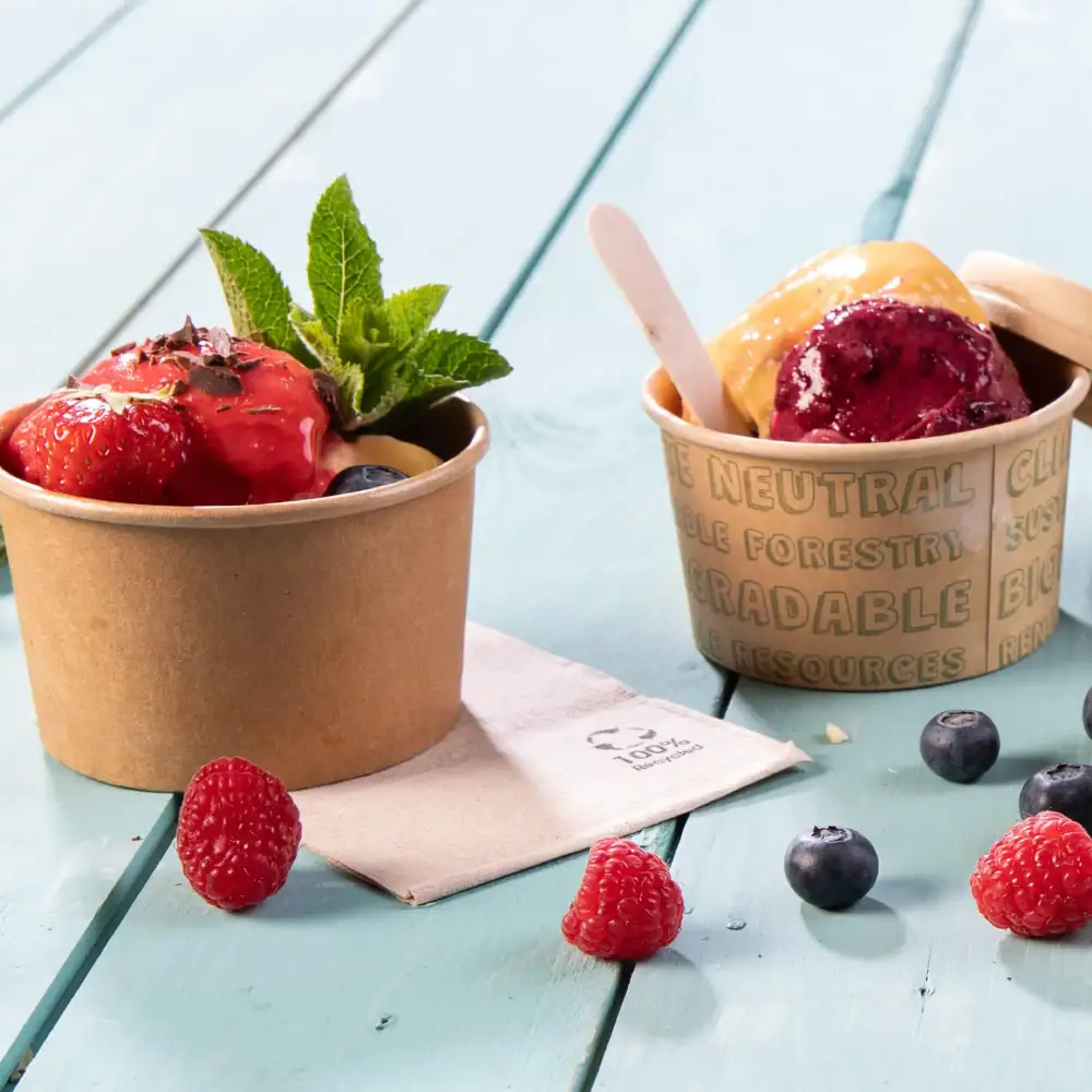Due coppe di gelato con frutti di bosco su una superficie in legno blu, una guarnita con fragole e menta, l'altra con gelato viola e giallo.