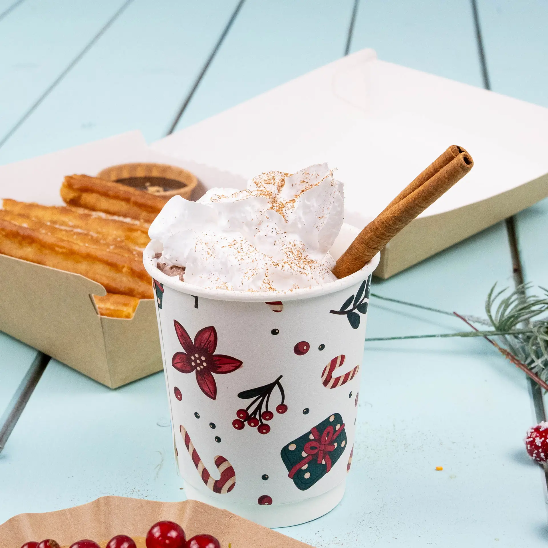 Bicchiere a doppia parete con design invernale, riempito di panna montata e un bastoncino di cannella, accanto a churros.