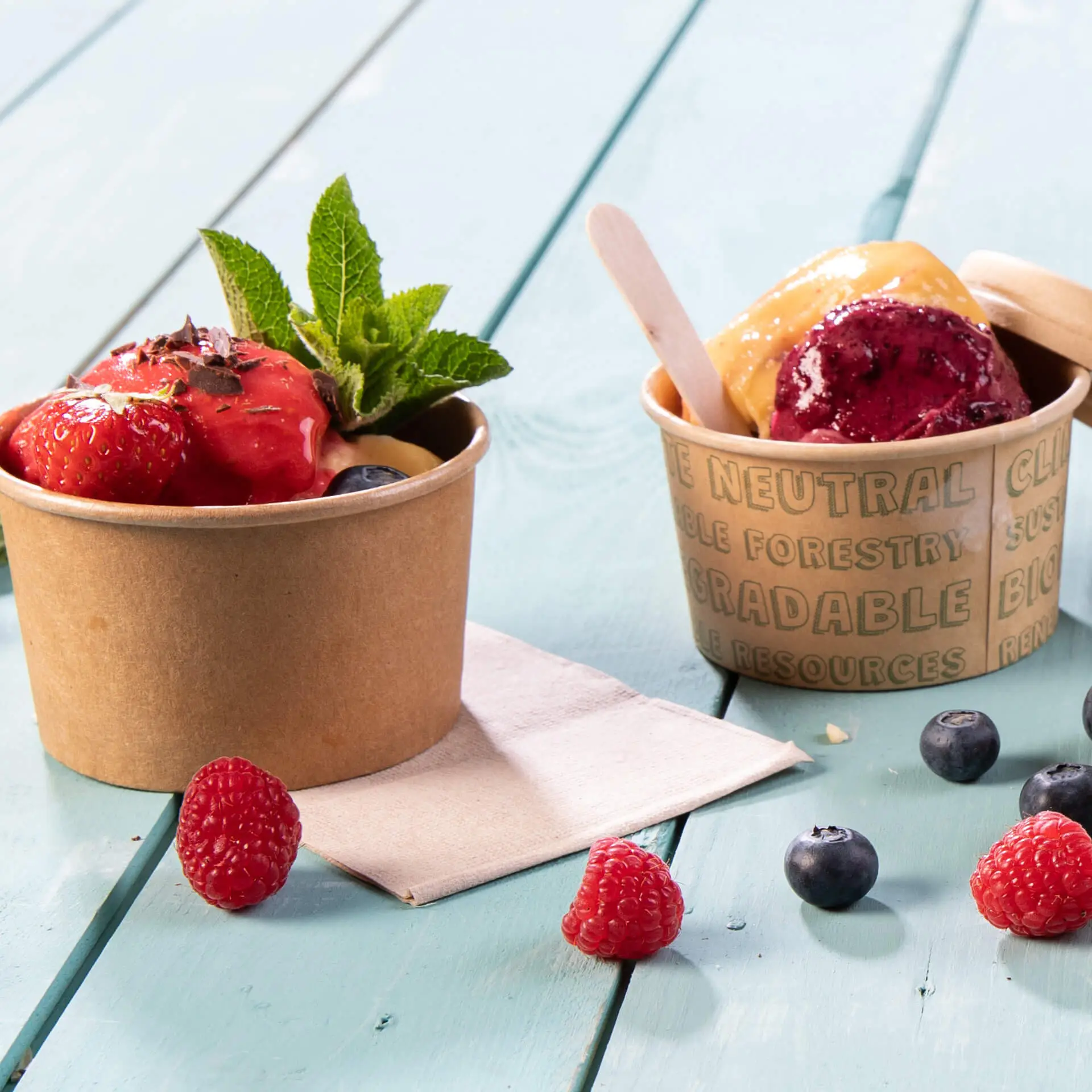 Due bicchieri di carta con gelato, uno guarnito con fragole e menta, e un tovagliolo su una superficie di legno blu.