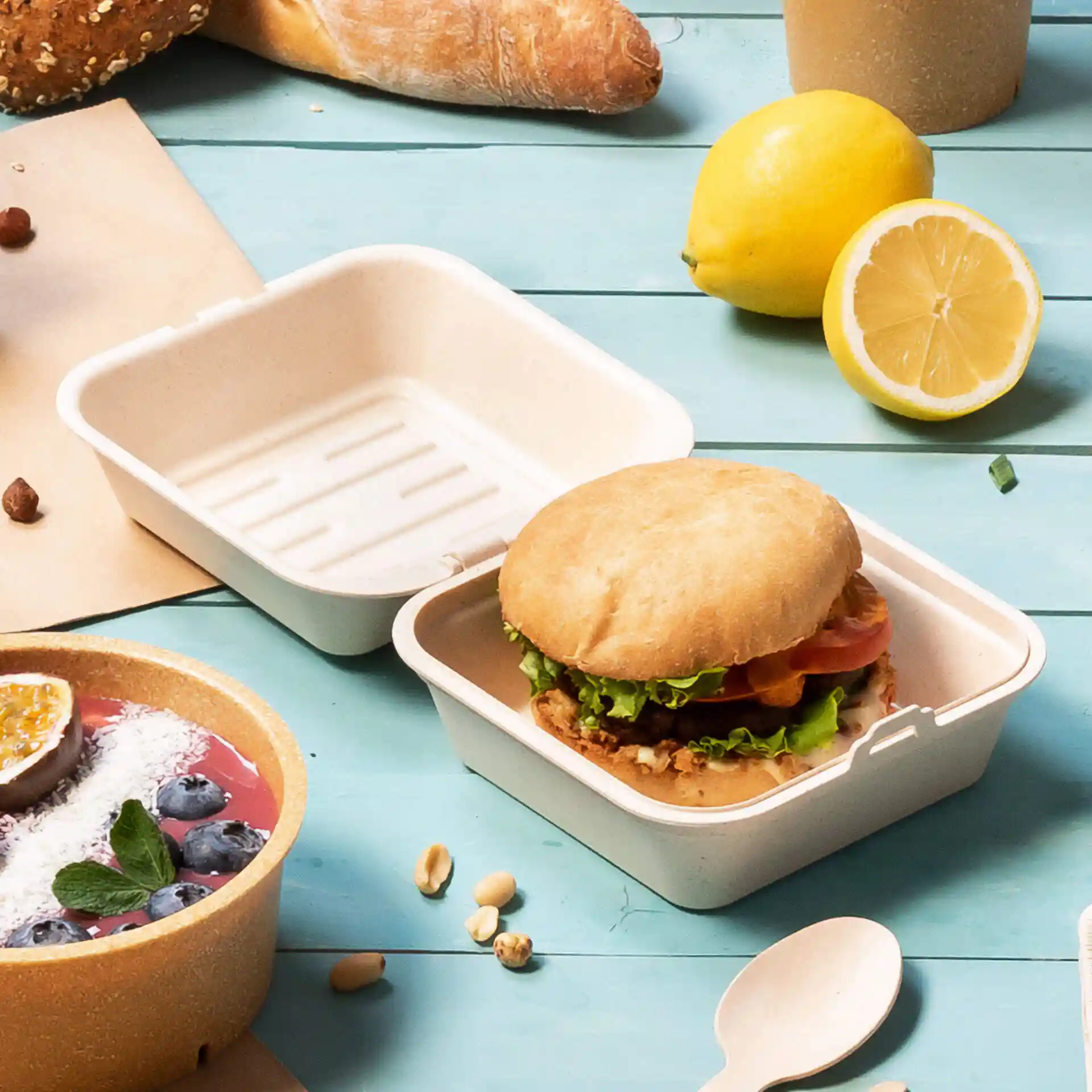 Scatola per hamburger riutilizzabile bianca cremosa con un panino, circondata da una ciotola di frutta e altri cibi.