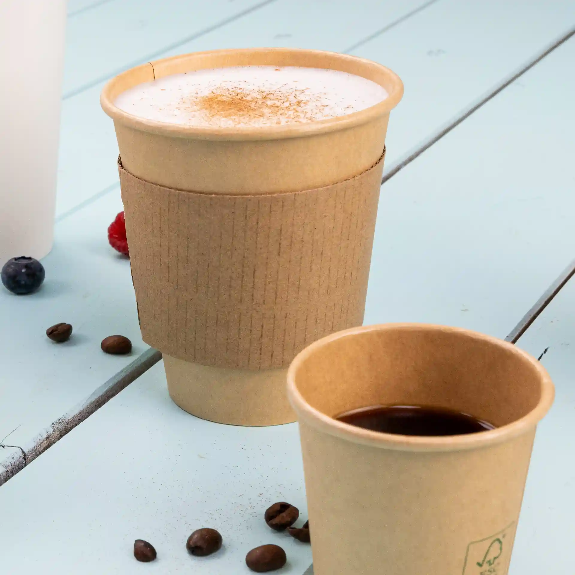 Bicchiere di carta marrone con manica di cartone, riempito con una bevanda schiumosa, accanto a una tazza di caffè più piccola e chicchi di caffè.