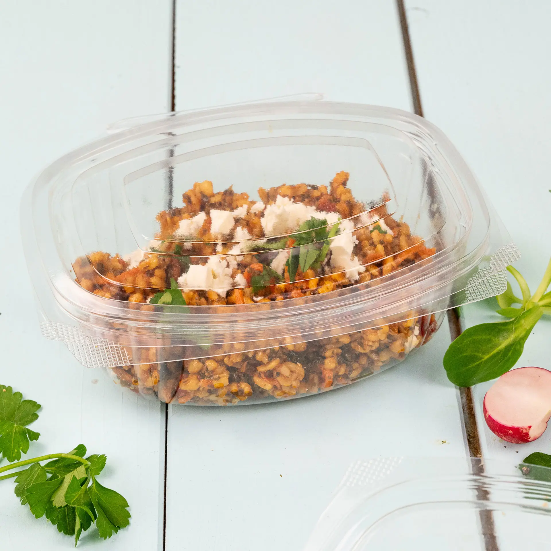 Ciotola Deli contenente un piatto freddo con verdure ed erbe, realizzata in plastica PLA biodegradabile, su un tavolo blu.