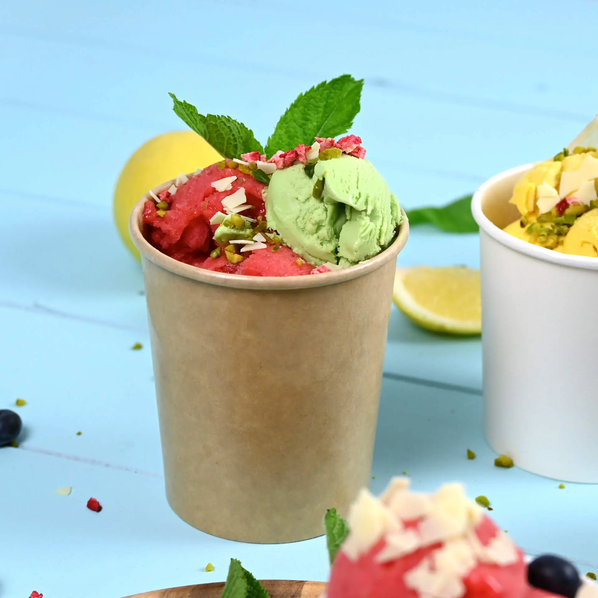 Grande coppetta di carta marrone piena di gelato rosa e verde, guarnita con noci e foglie di menta, su sfondo azzurro chiaro.