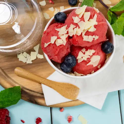 Ciotola di sorbetto ai lamponi guarnita con mirtilli e scaglie di cioccolato bianco, cucchiaio di legno e foglie di menta su una superficie di legno.