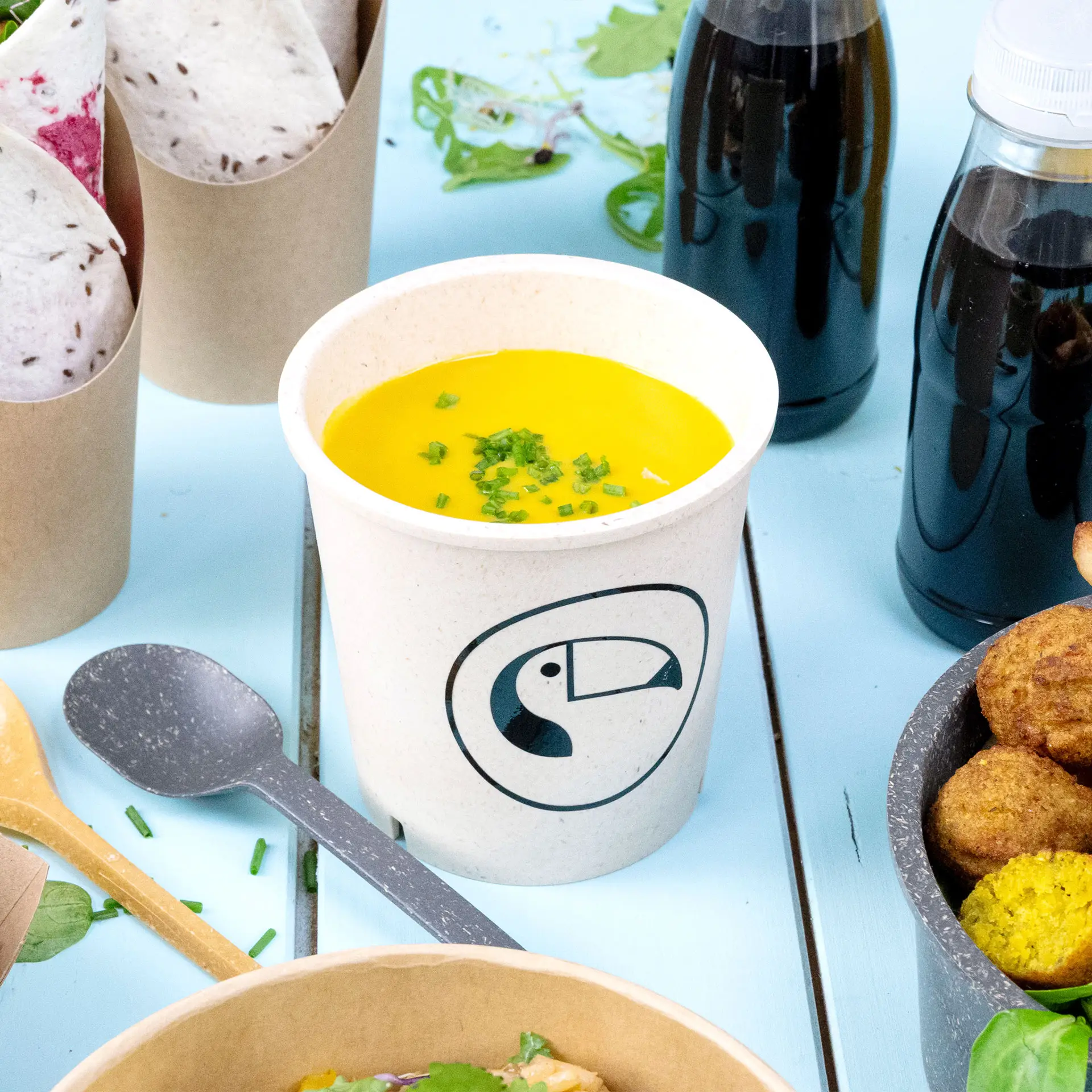 Una tazza da zuppa riutilizzabile bianco crema, piena di zuppa gialla e guarnita con erba cipollina tritata; cucchiaio accanto.