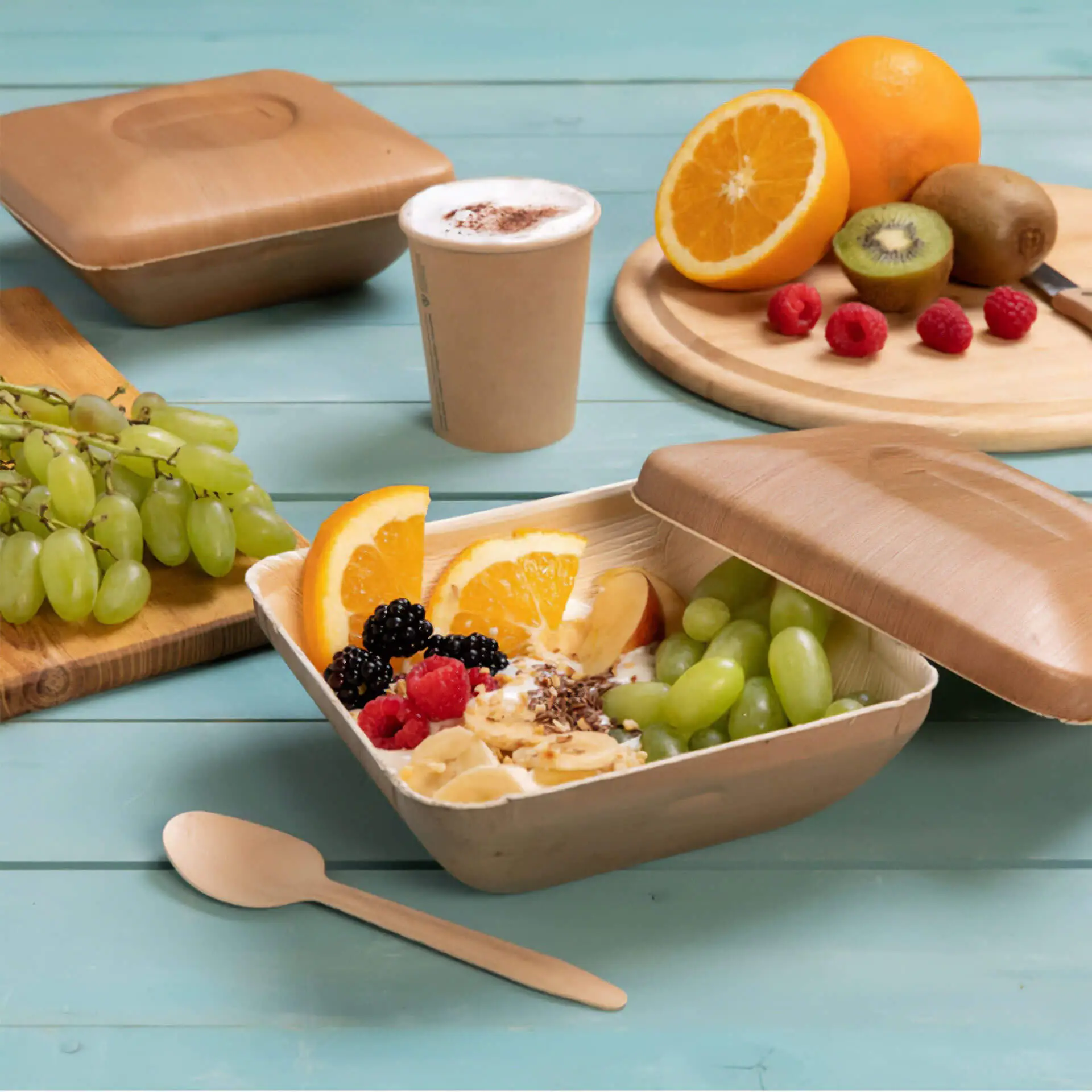 Una ciotola quadrata in foglia di palma con coperchio riempita di frutta; include uva verde, fette d'arancia, lamponi e more.