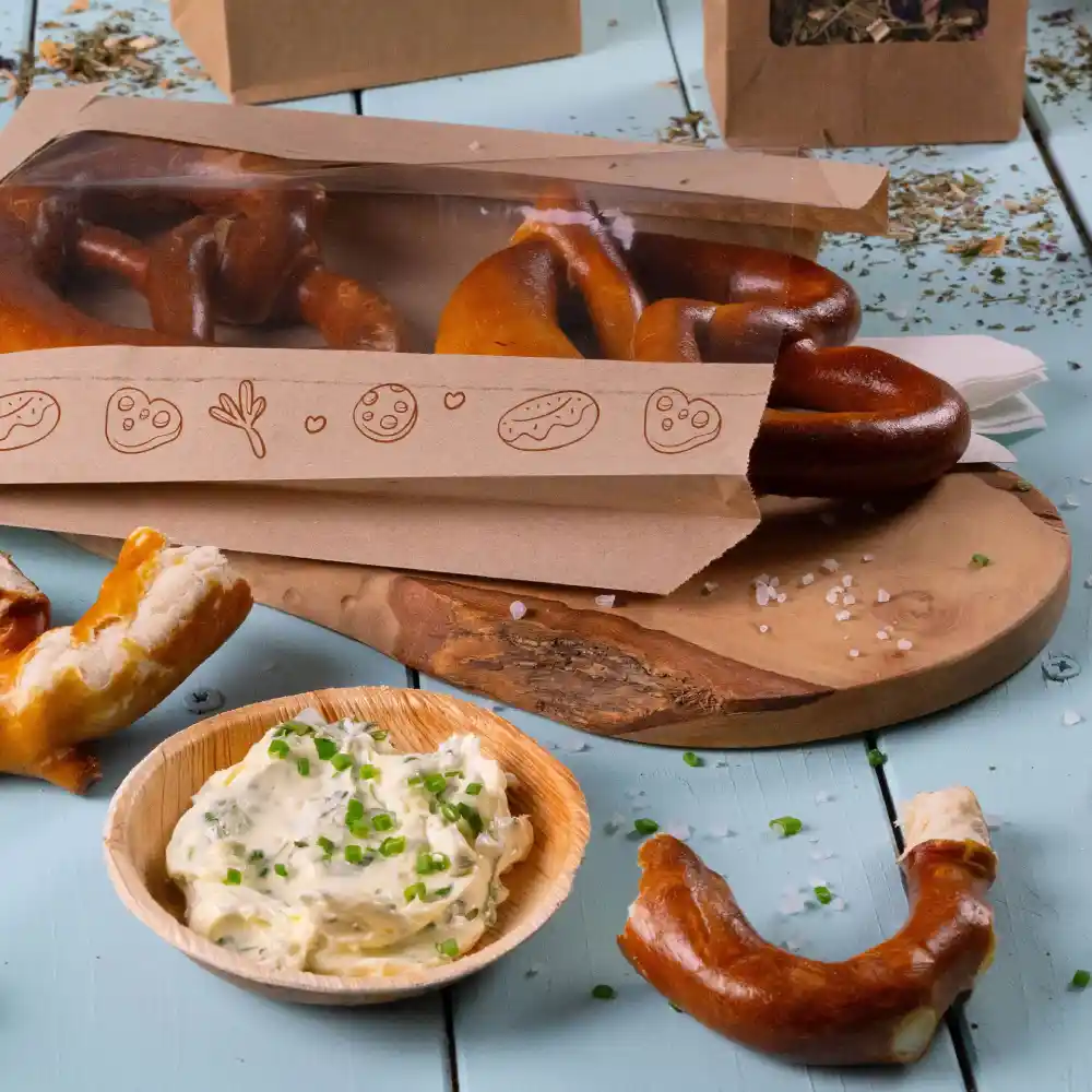 Pretzel in un sac trasparente con un nastro decorativo, servite con una ciotola di salsa cremosa e erbe verdi su un tagliere di legno.