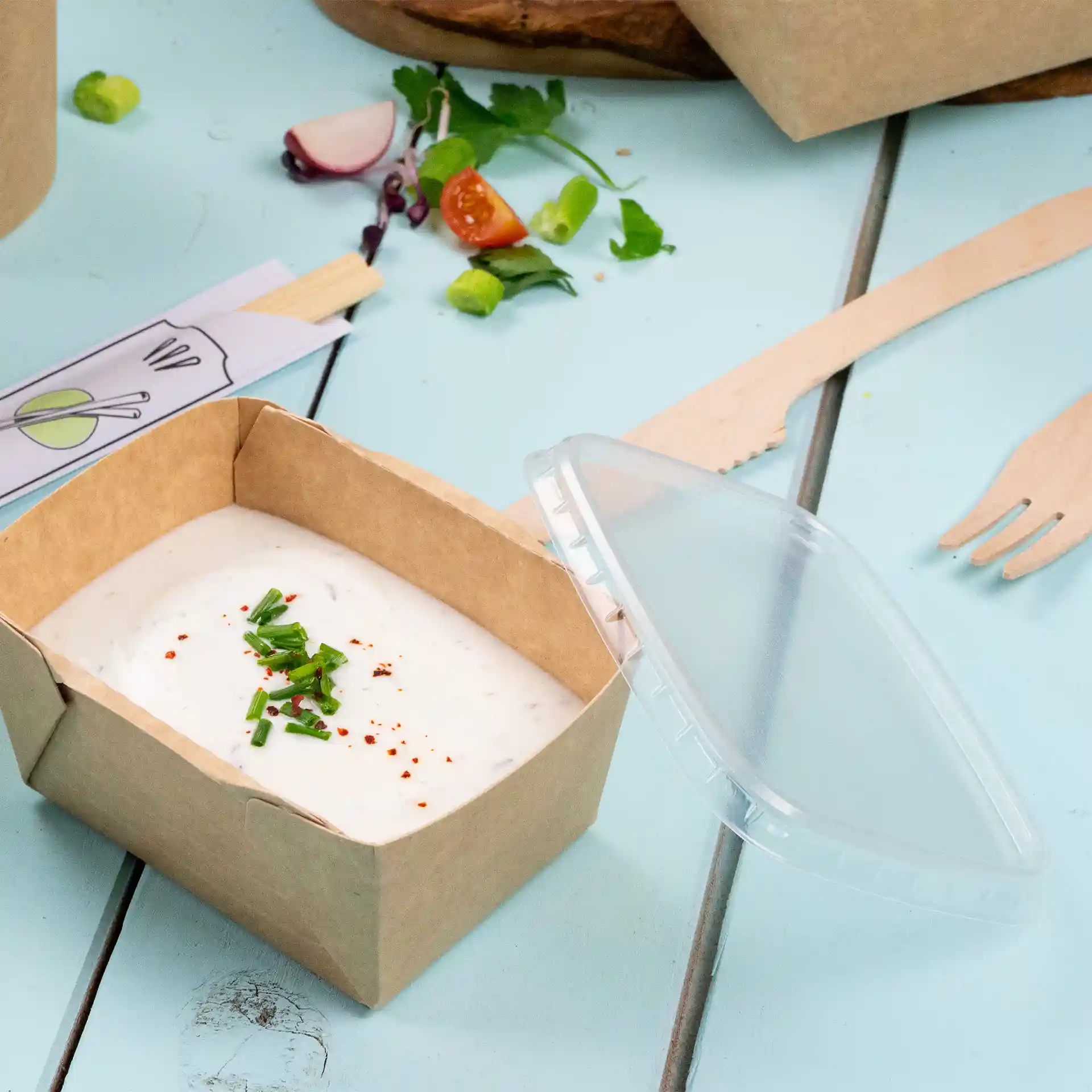 Contenitore rettangolare in cartone con coperchio trasparente in rPET, contenente una salsa cremosa guarnita con erba cipollina tritata e fiocchi di peperoncino rosso.