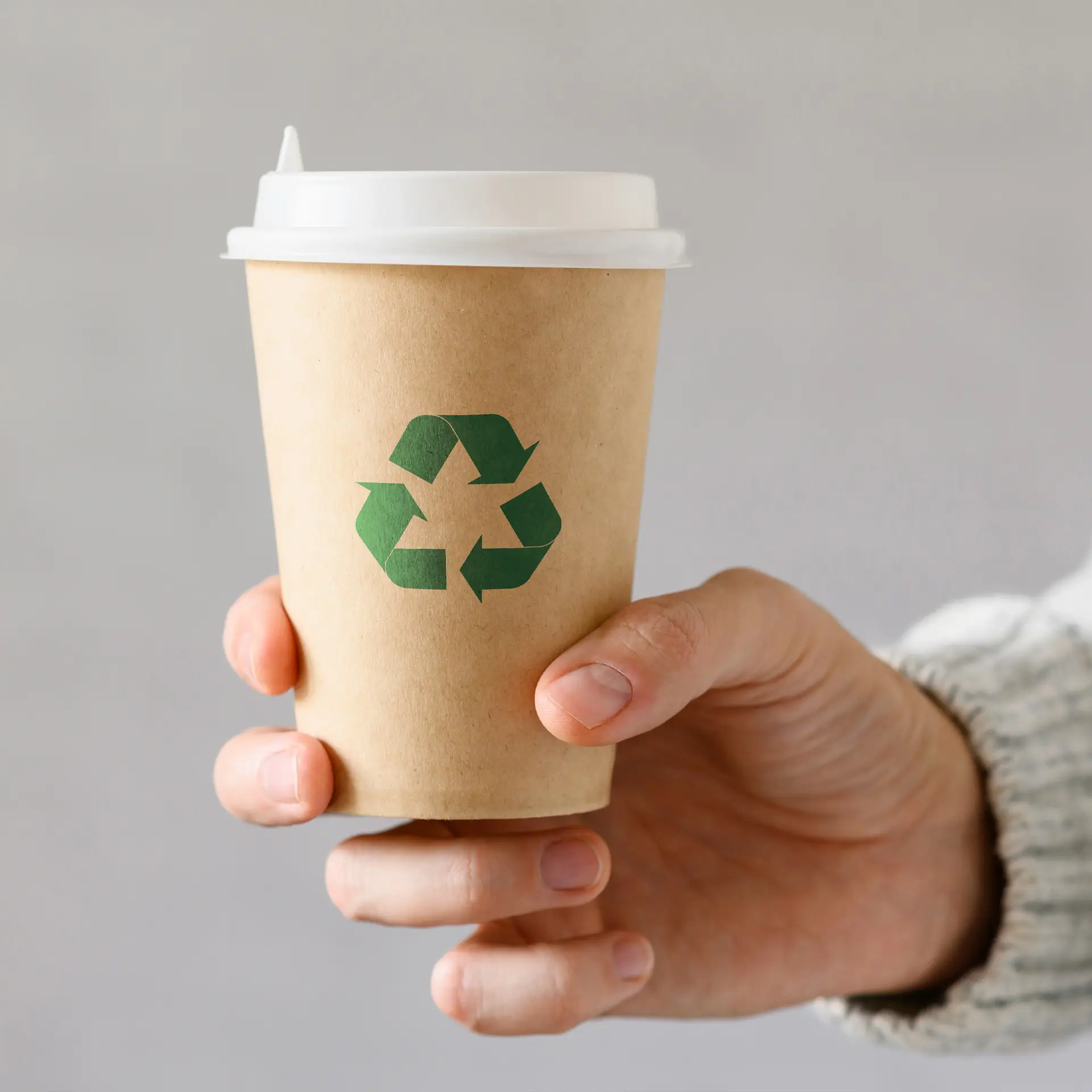 Una tazza di carta marrone con un logo di riciclaggio verde, tenuta da una mano, su uno sfondo neutro.