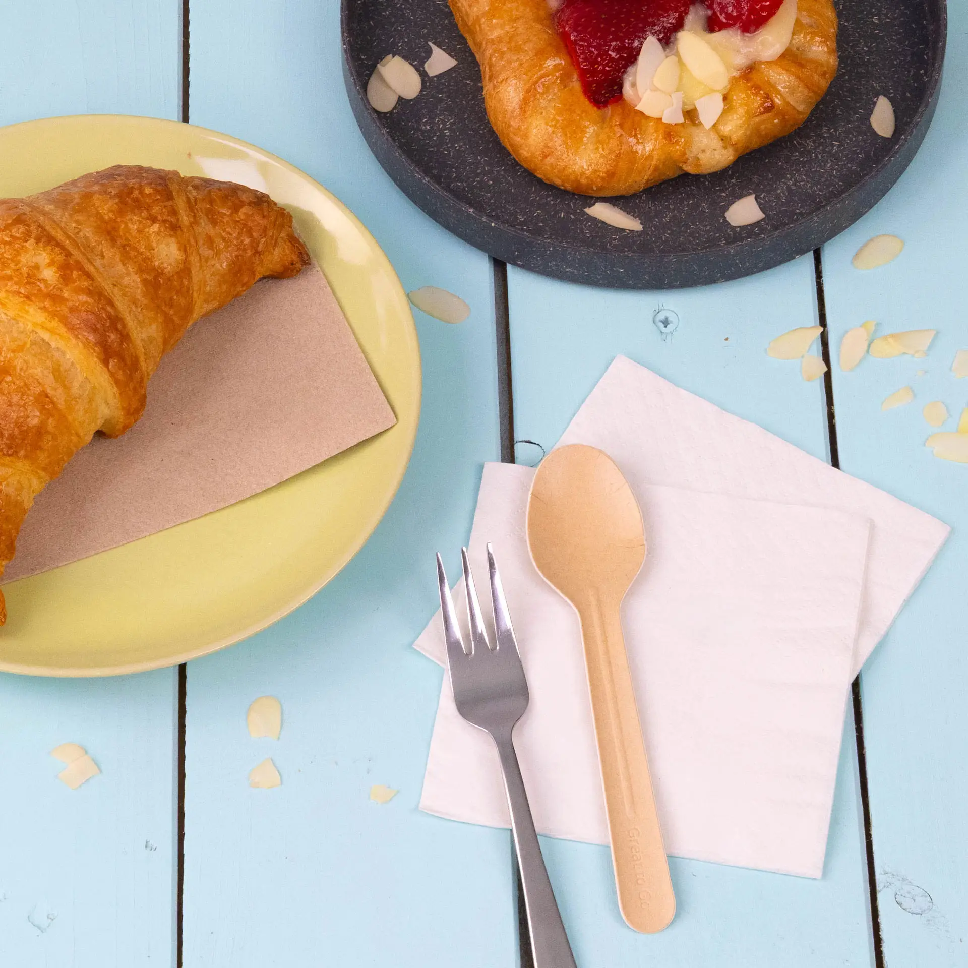 Immagine di un croissant su un piatto giallo, un dolce su un piatto scuro, un cucchiaio di legno, una forchetta d'argento e tovaglioli bianchi.