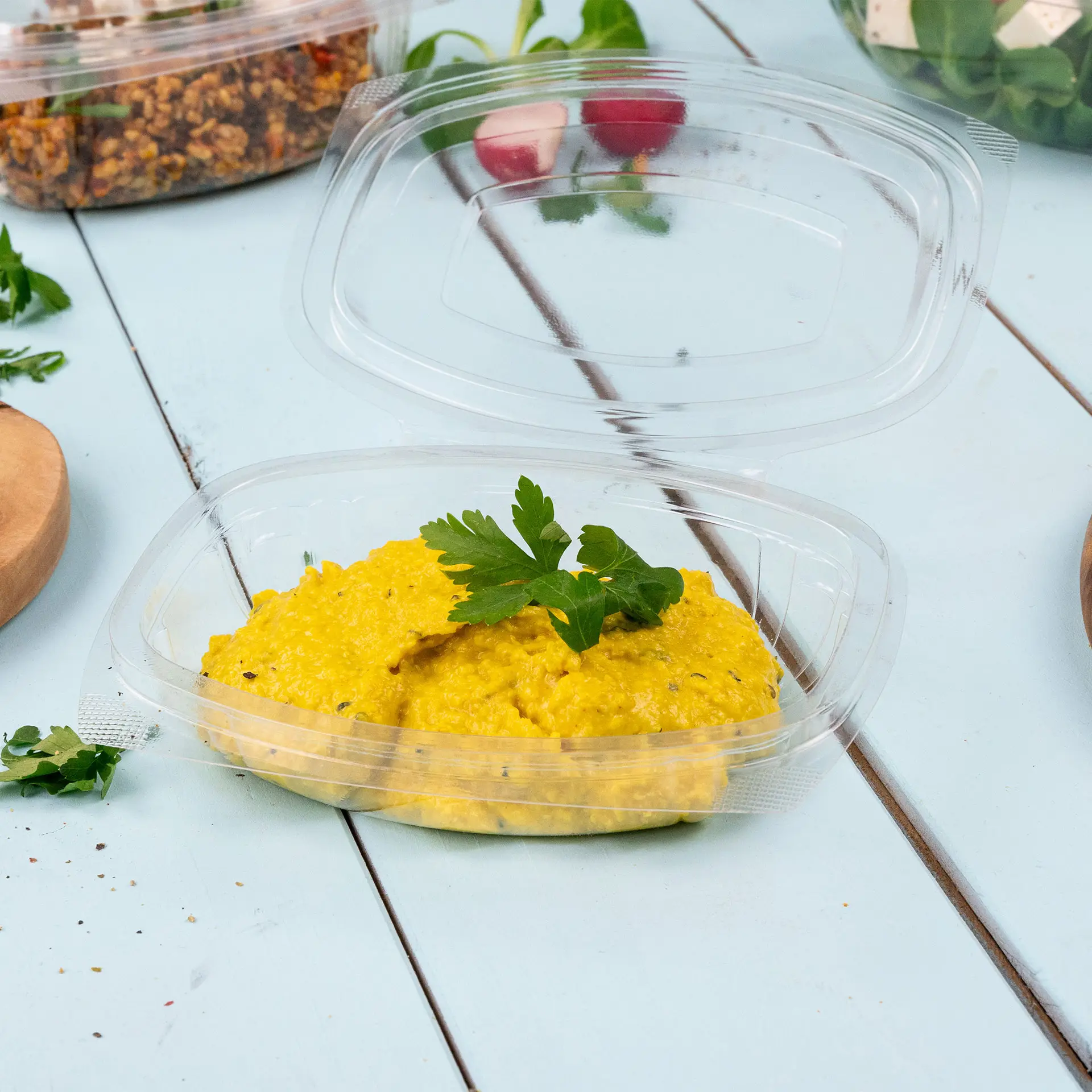 Ciotola deli con salsa gialla, guarnita con prezzemolo; coperchio trasparente parzialmente sollevato, circondato da ingredienti per insalata.