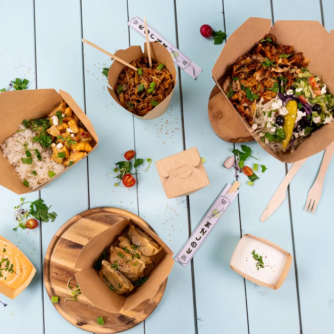 Contenitori per cibo da asporto assortiti con noodles, riso, insalata e ravioli su una superficie in legno blu.