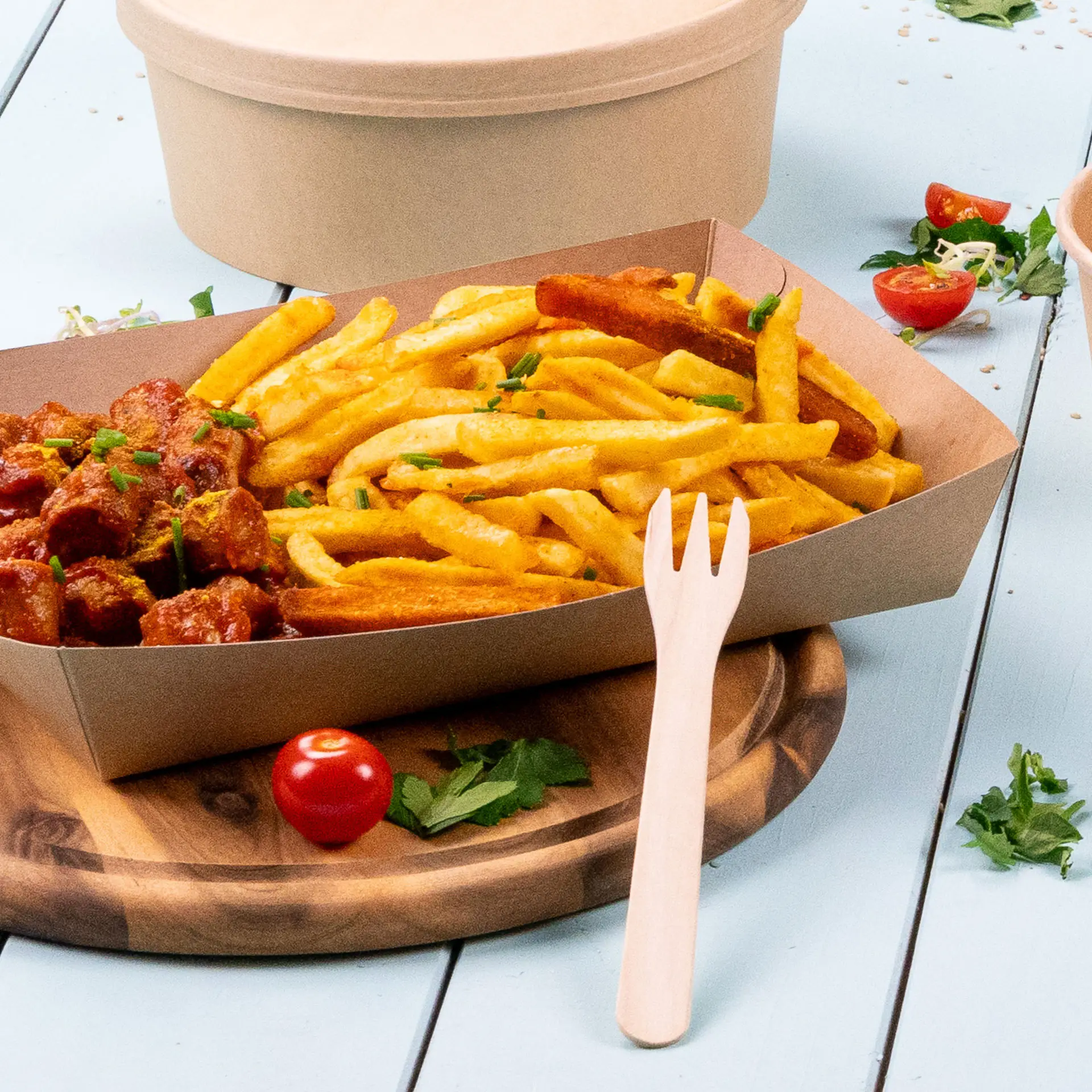 Una forchetta in legno biodegradabile accanto a una porzione di patatine e a un piatto di carne, su un tagliere di legno.