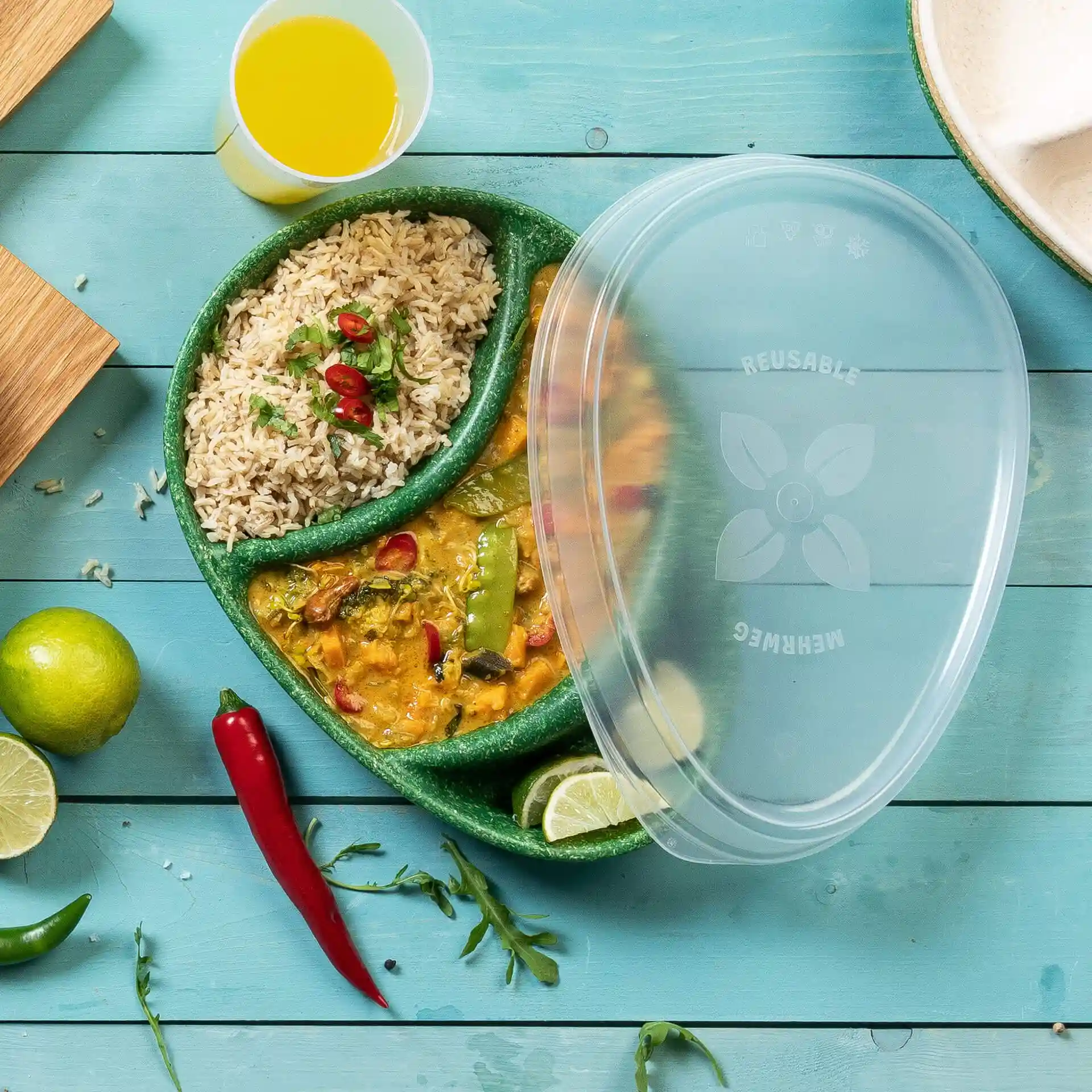 Coperchio riutilizzabile trasparente su un piatto diviso contenente riso e curry, con lime e peperoncini su una superficie in legno blu.