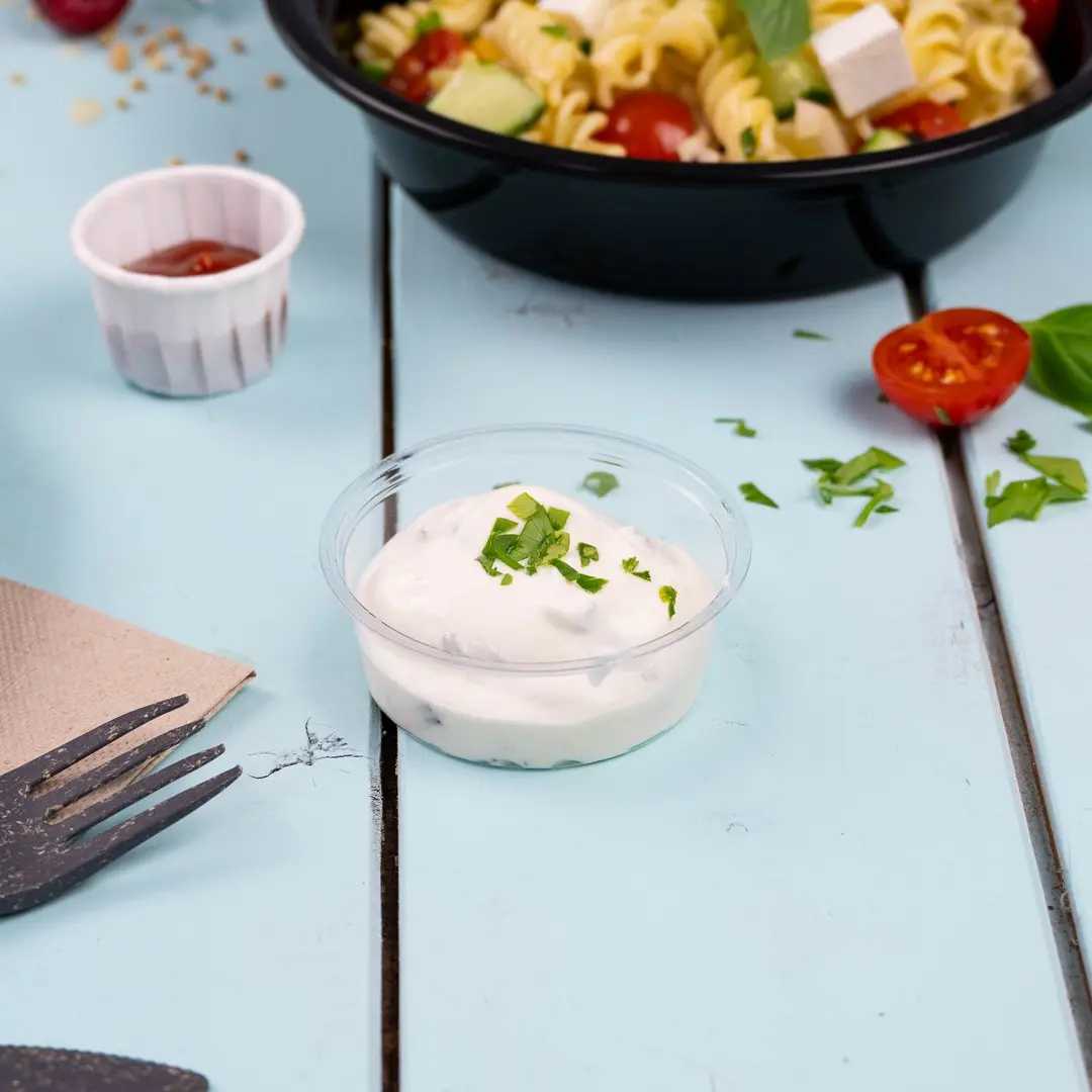 Bicchiere di plastica trasparente con salsa cremosa guarnita con erbe tritate, posato su una superficie in legno blu chiaro.