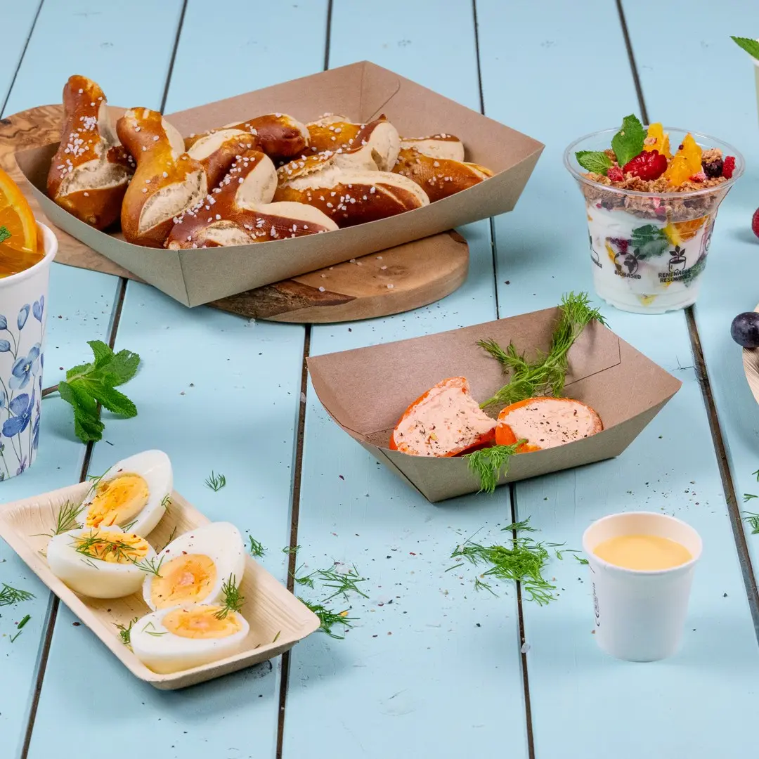 Snack assortiti tra cui pretzel, uova sode, parfait di yogurt e un piccolo bicchiere di salsa su una superficie di legno blu.