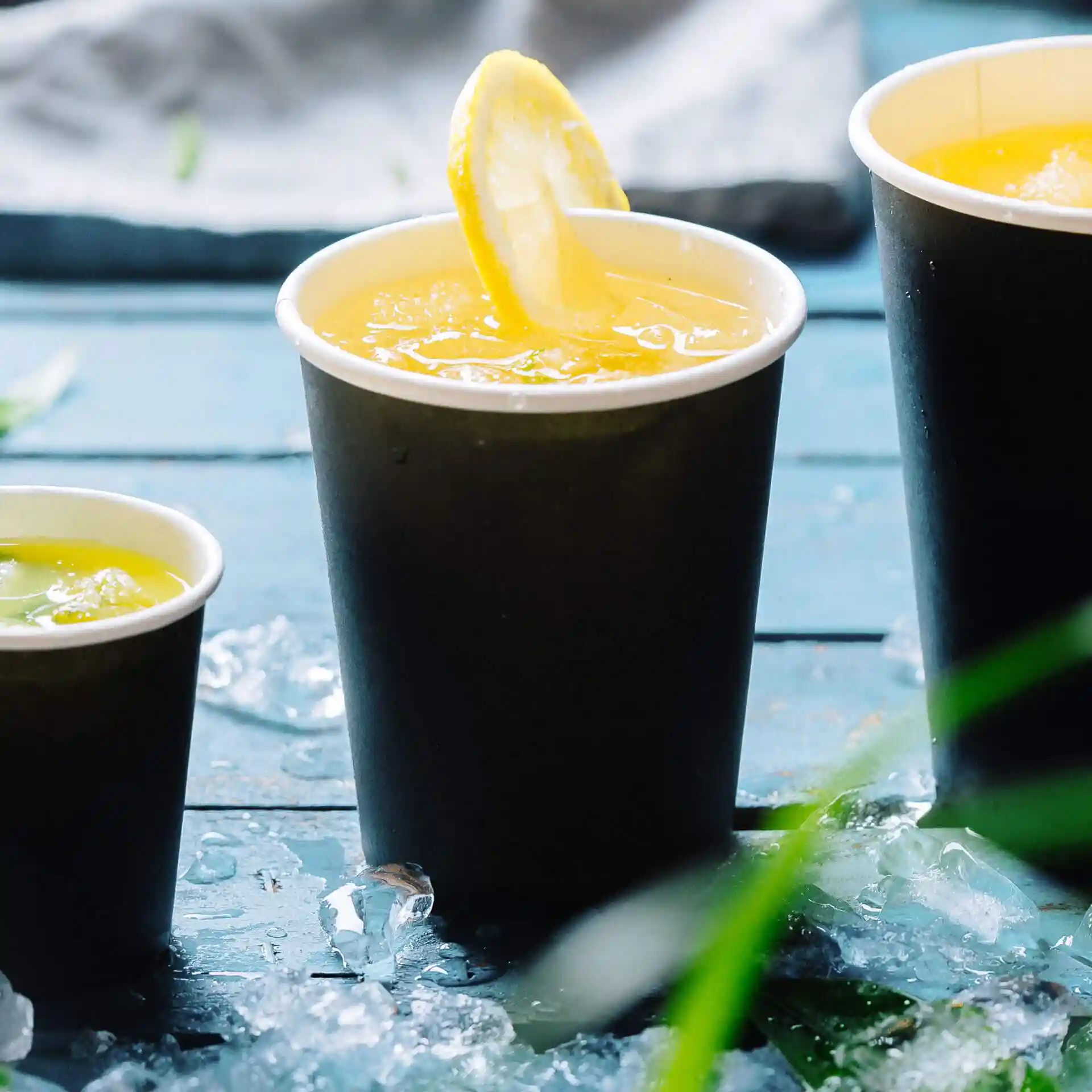 Tre bicchieri di carta neri pieni di bevande, uno guarnito con una fetta di limone, su una superficie di legno blu con ghiaccio.