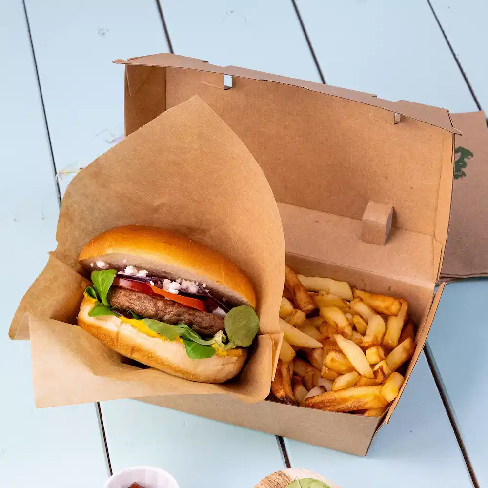 Burger con condimenti in una scatola da asporto accanto a una porzione di patatine fritte.