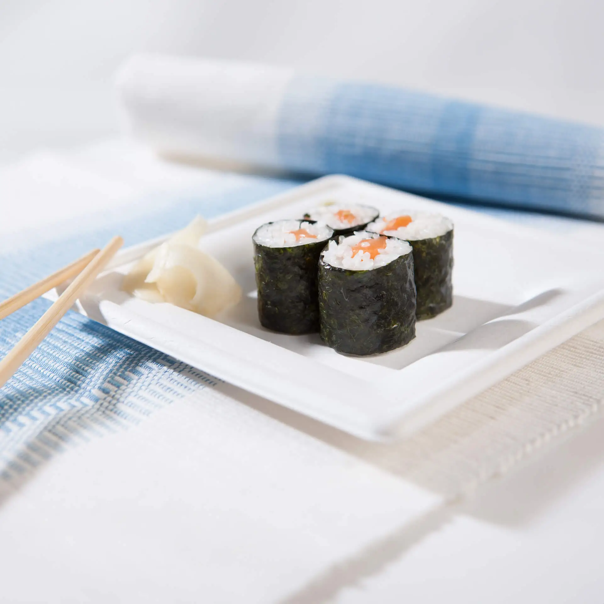 Piatto quadrato in canna da zucchero con rotoli di sushi e zenzero marinato, su una mise en place blu e bianca.